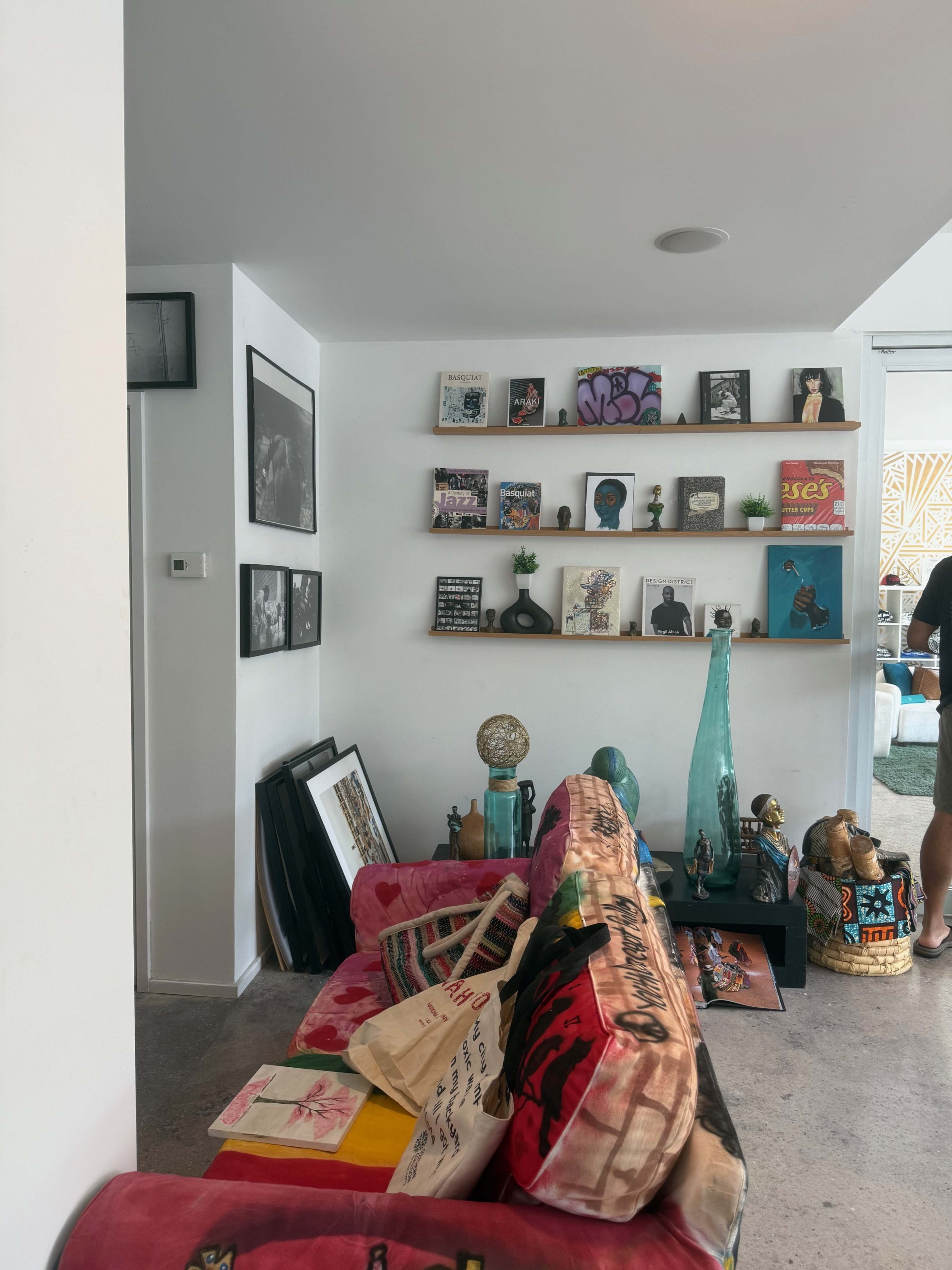 The image shows a living room corner featuring a pink couch, a shelf with various books and decorative items, and some framed pictures on the wall.