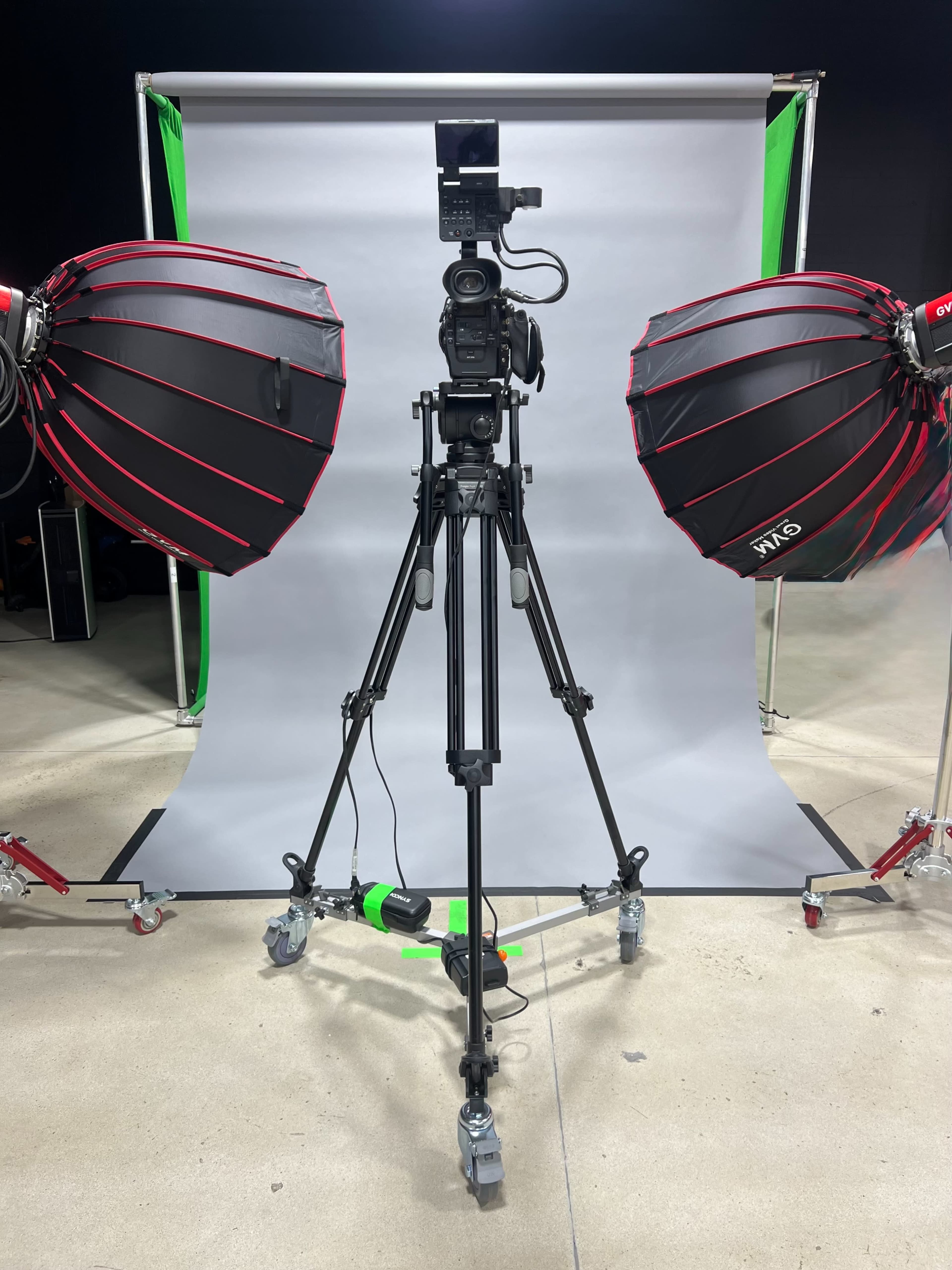 The image shows a camera mounted on a tripod, flanked by two large softbox lights, in front of a gray backdrop.