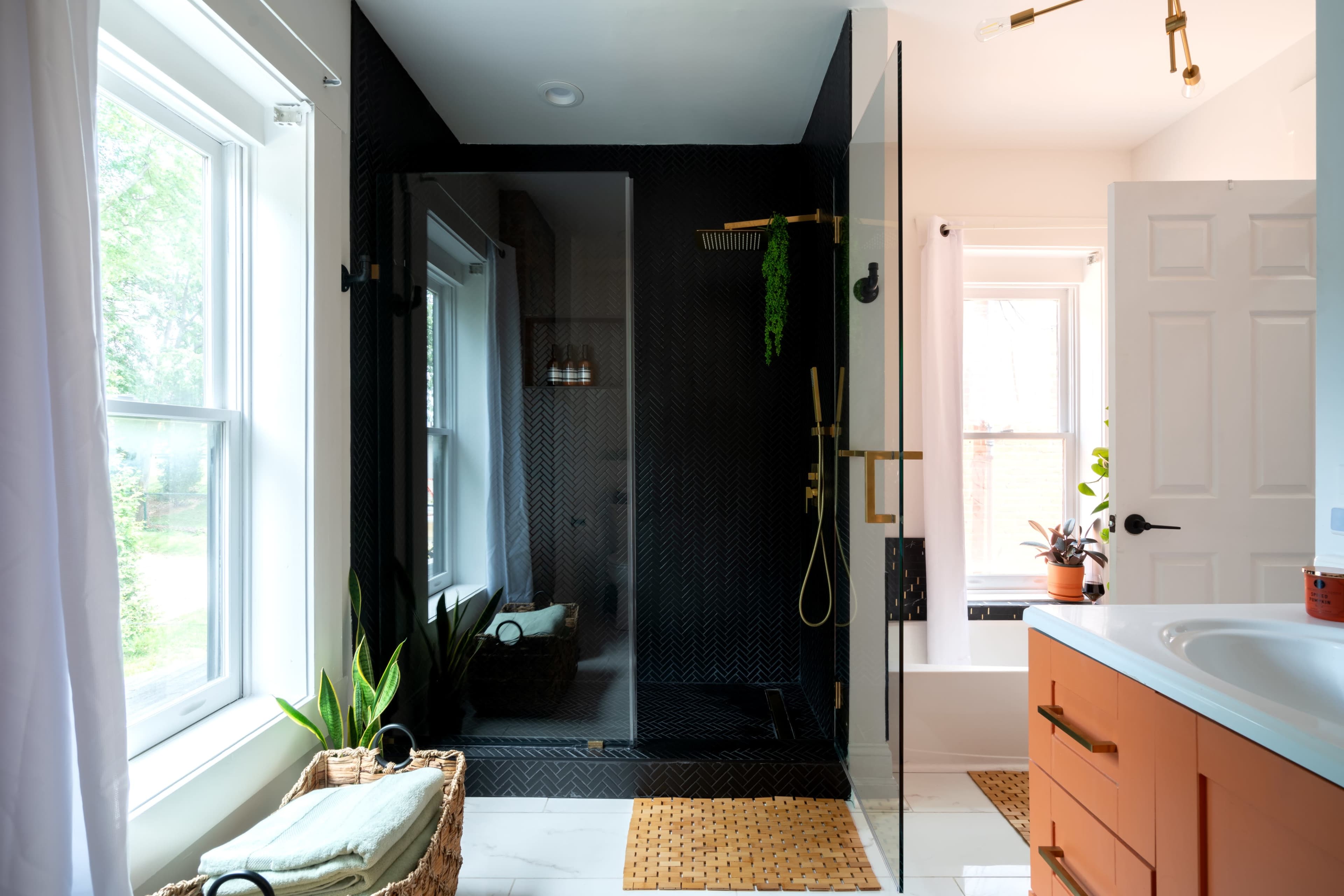 A modern bathroom featuring a black tiled shower with glass doors, a light-colored sink countertop, and plants near the window.