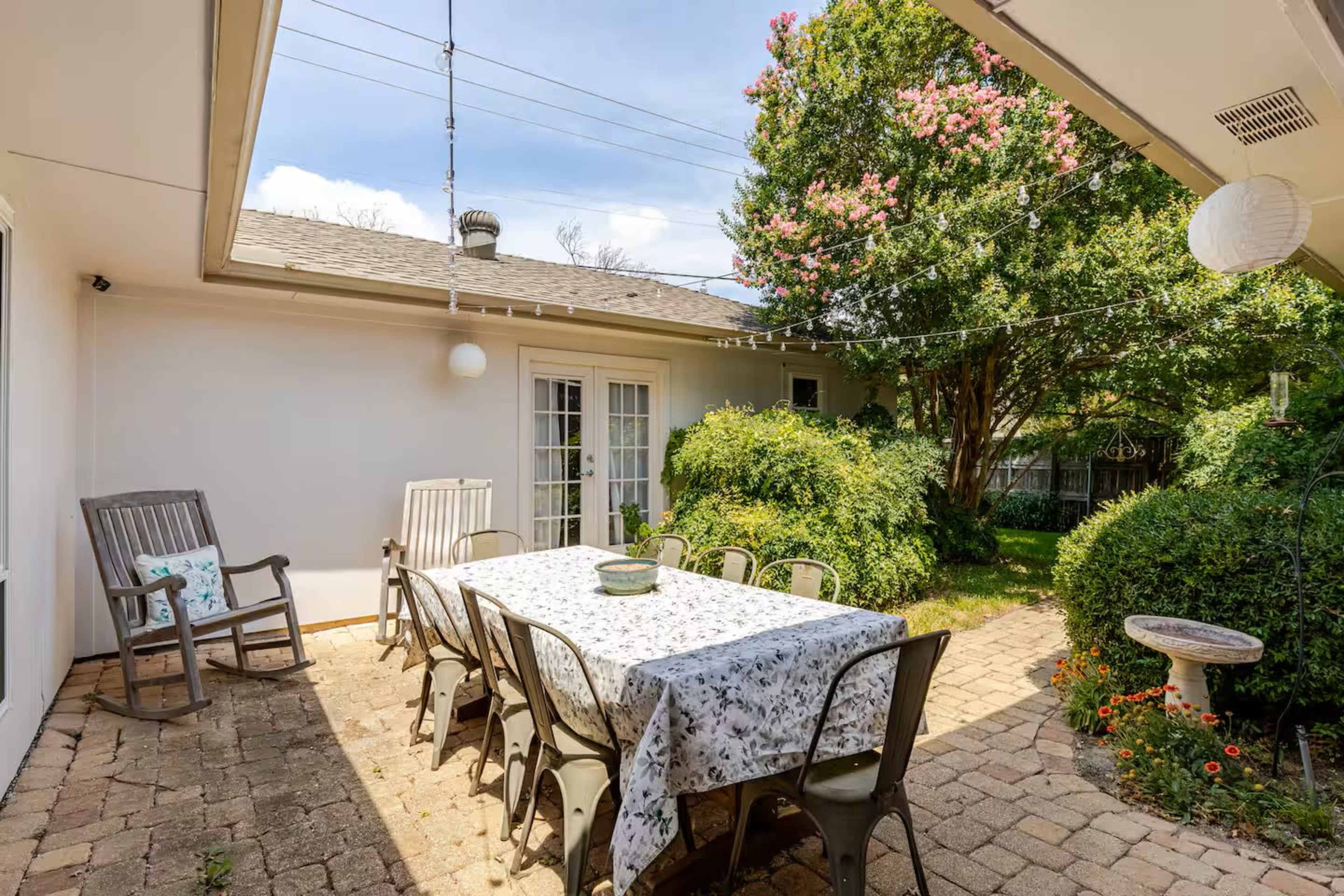 The image shows an outdoor patio with a large table surrounded by chairs, bordered by greenery and adorned with string lights.