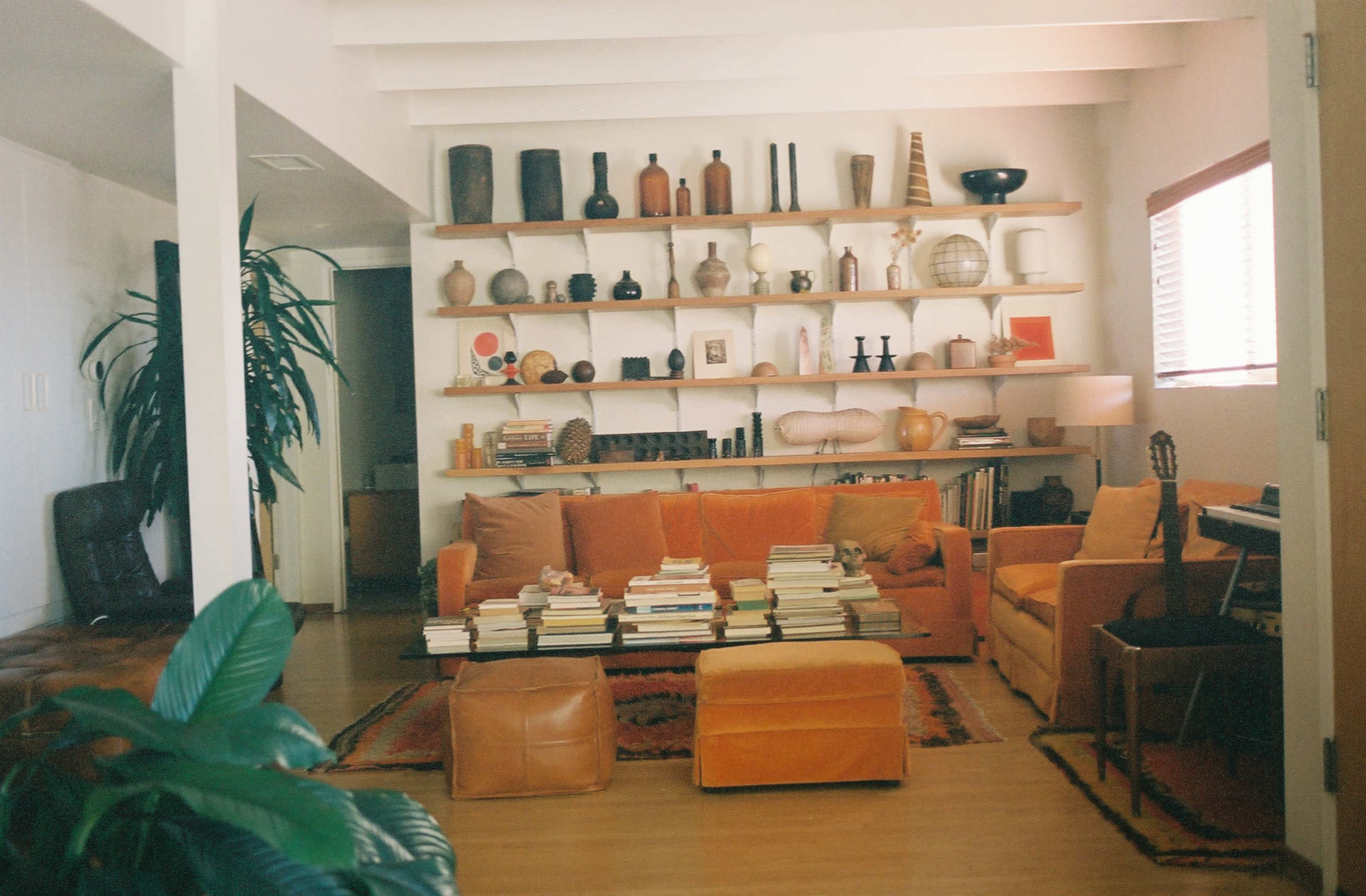A cozy living room with an orange sofa, a coffee table stacked with books, and shelves adorned with various decorative items against a bright wall.