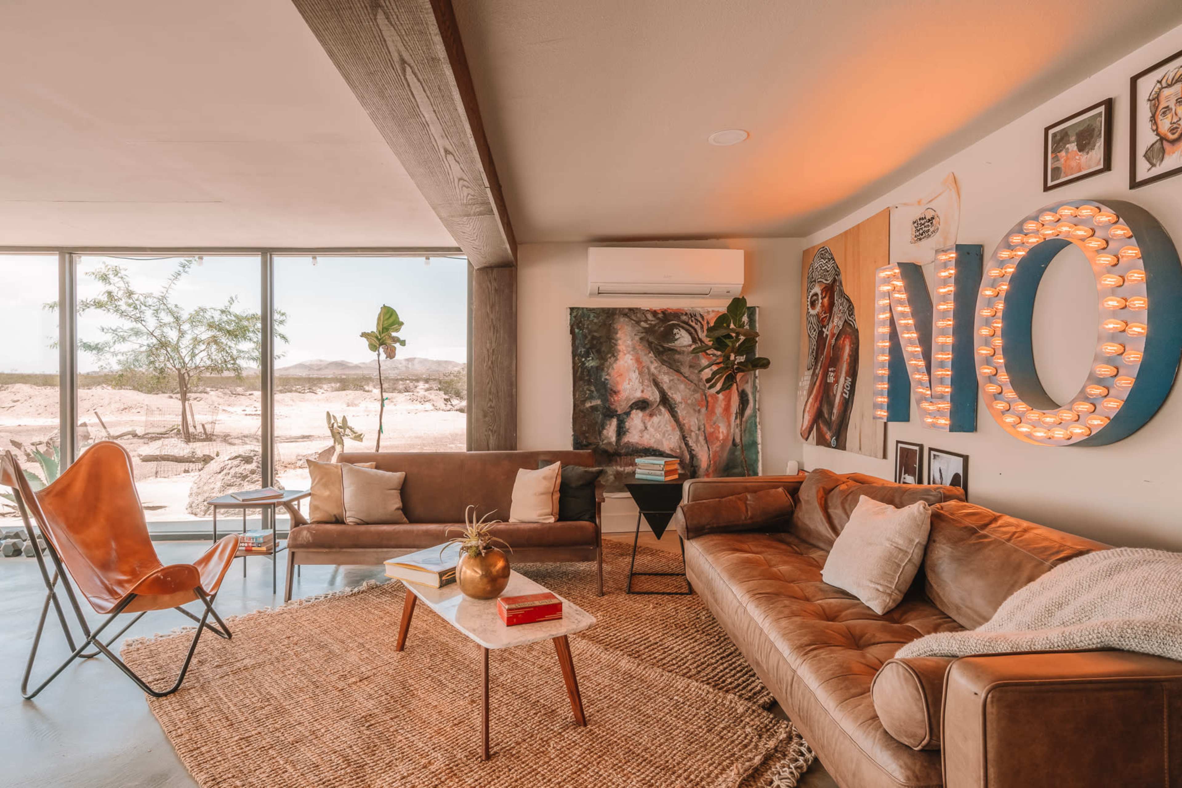 The living room features a large window with a view of a desert landscape, a leather sofa, a butterfly chair, a coffee table, and artwork on the walls.