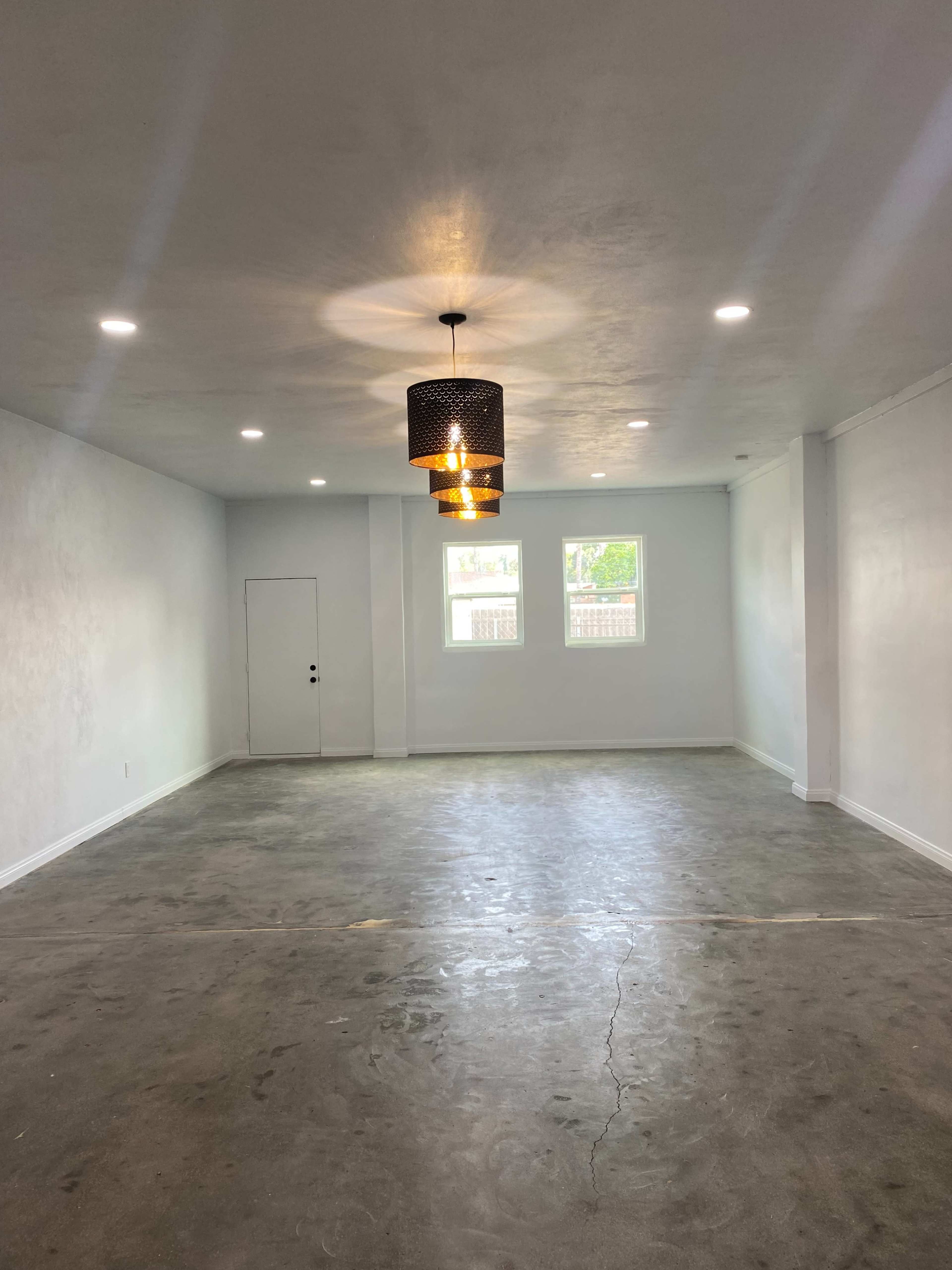 The image shows an empty room with a concrete floor, white walls, and a decorative light fixture hanging from the ceiling.