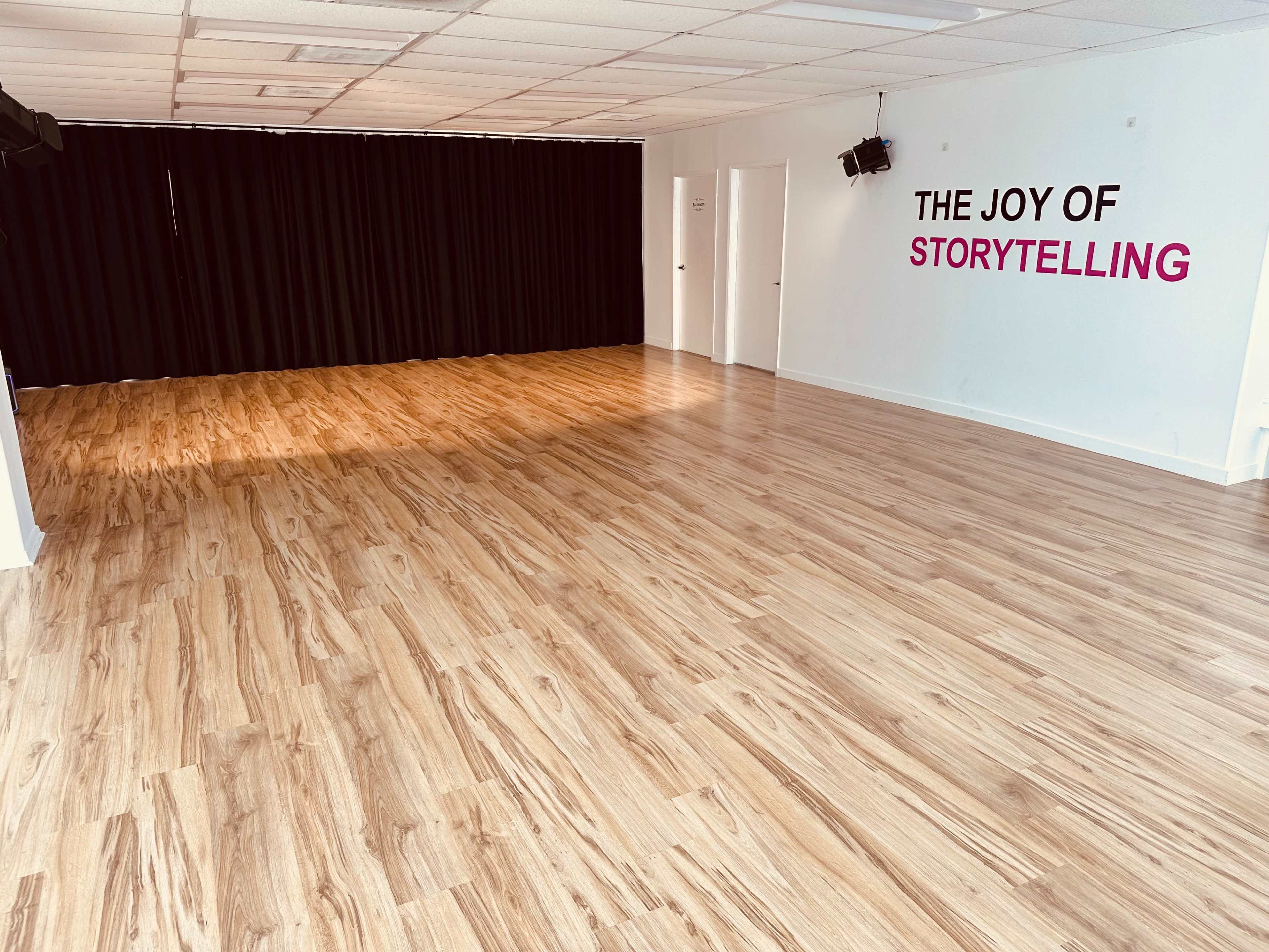 A spacious, empty room with wooden flooring and a black curtain at one end, featuring the phrase "THE JOY OF STORYTELLING" on the wall.