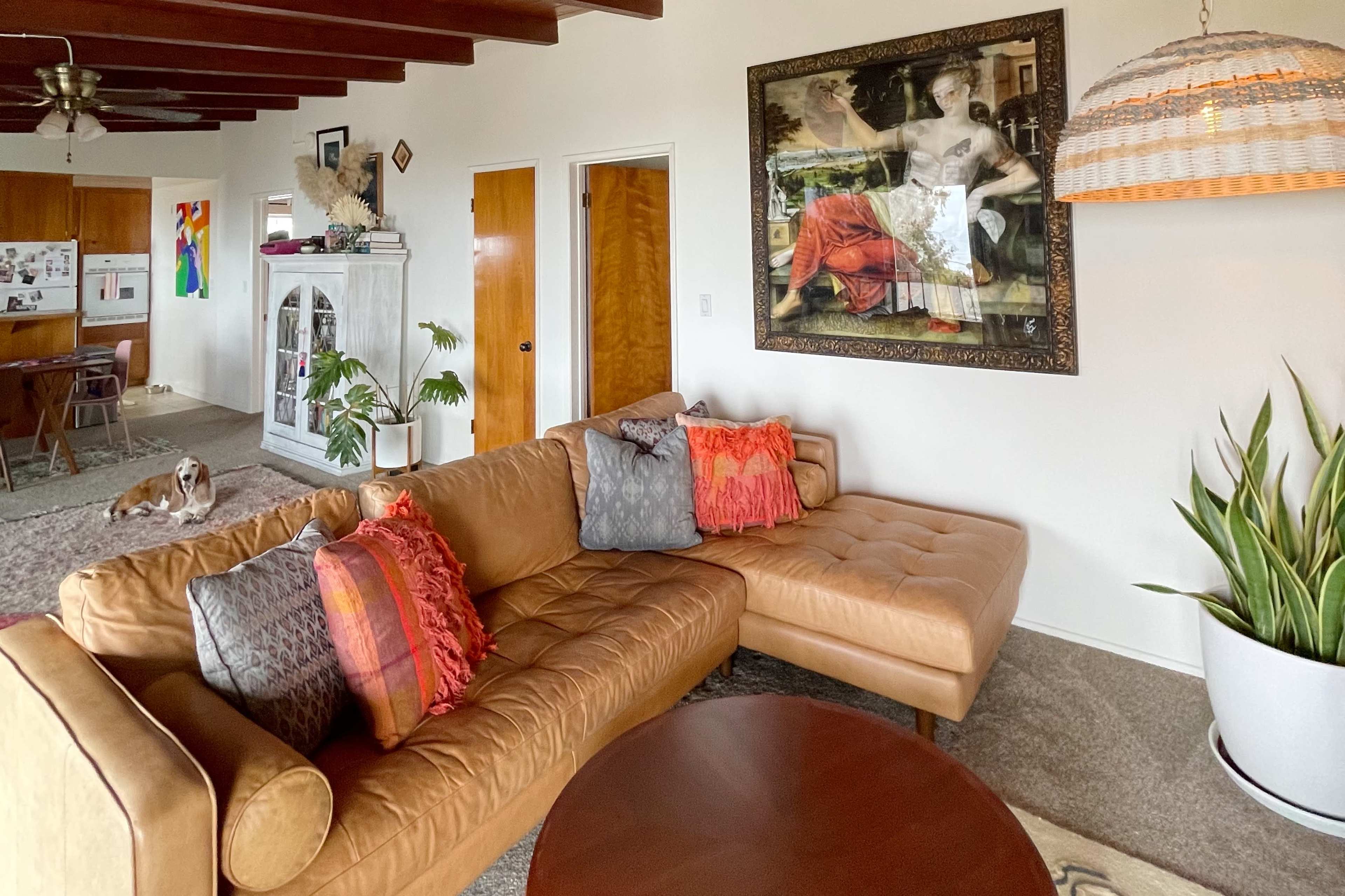 The living room features a tan sectional sofa, a round wooden coffee table, a large painting on the wall, and a small dog lying on the carpet.