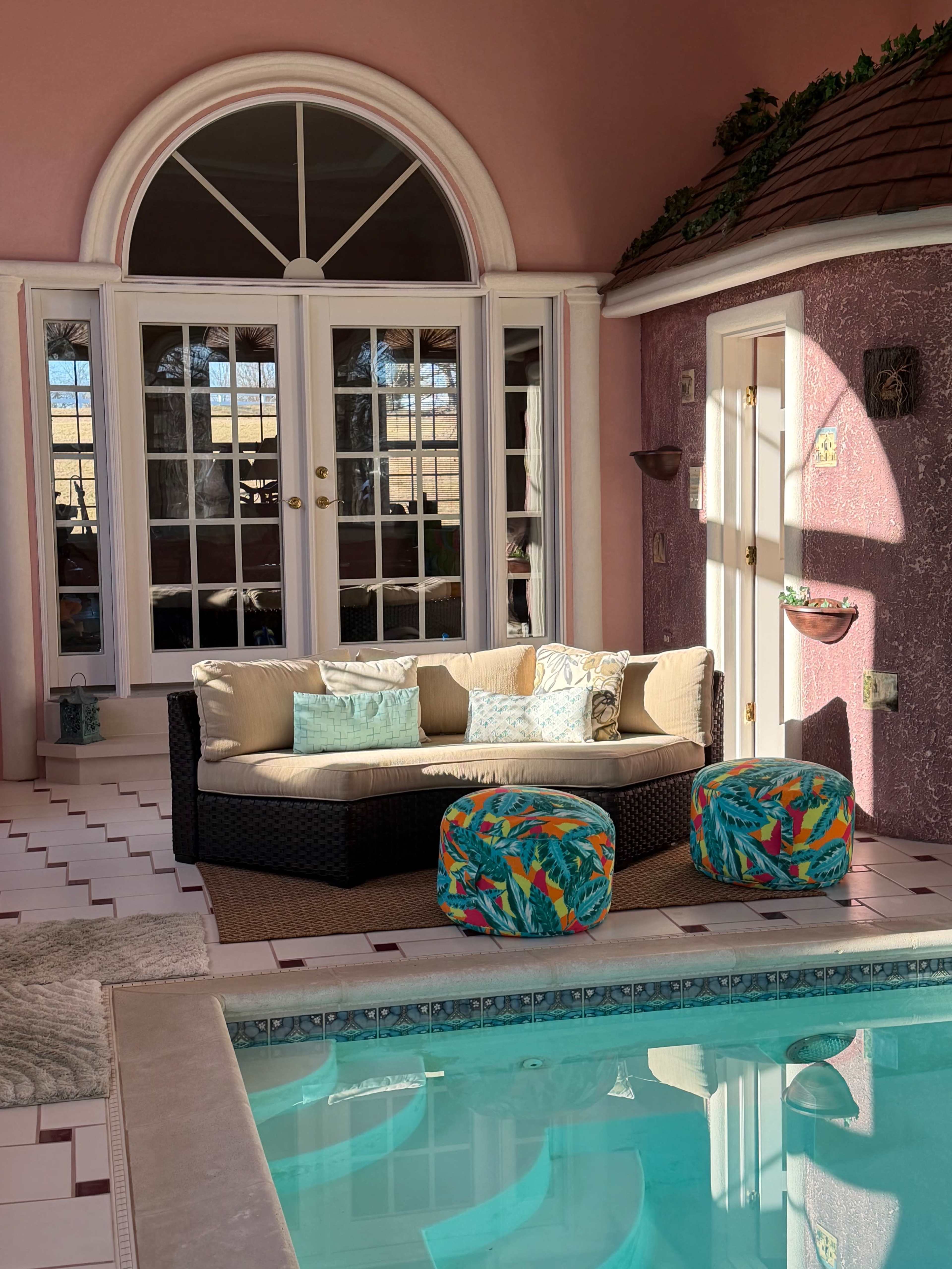The image shows a cozy indoor area with a pool, featuring a wicker sofa with cushions and colorful poufs beside it, all surrounded by large windows and pink walls.