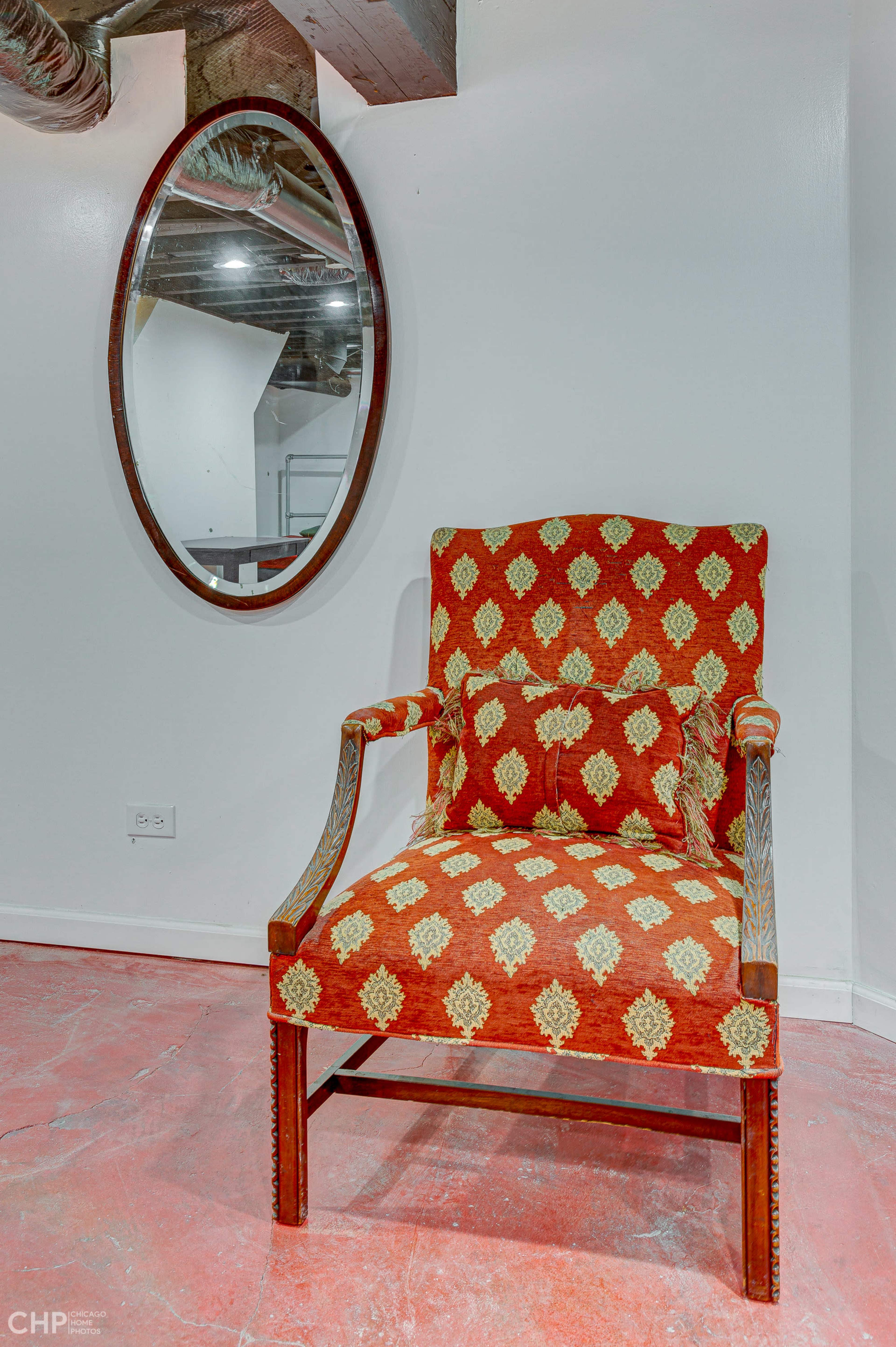 A red-patterned upholstered chair with a matching decorative pillow positioned in front of an oval mirror on a white wall.