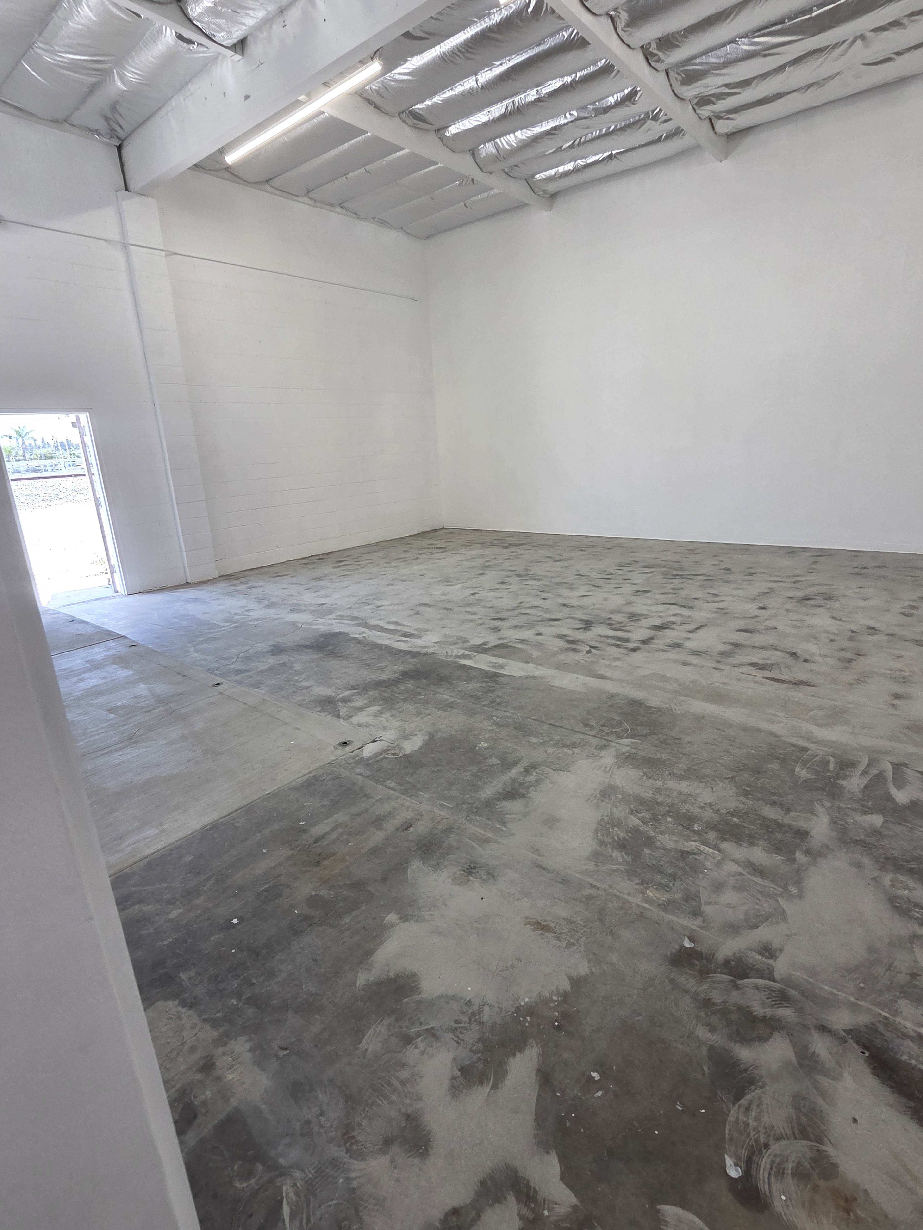 The image shows an empty, spacious interior of a commercial building with unfinished concrete flooring and white walls.