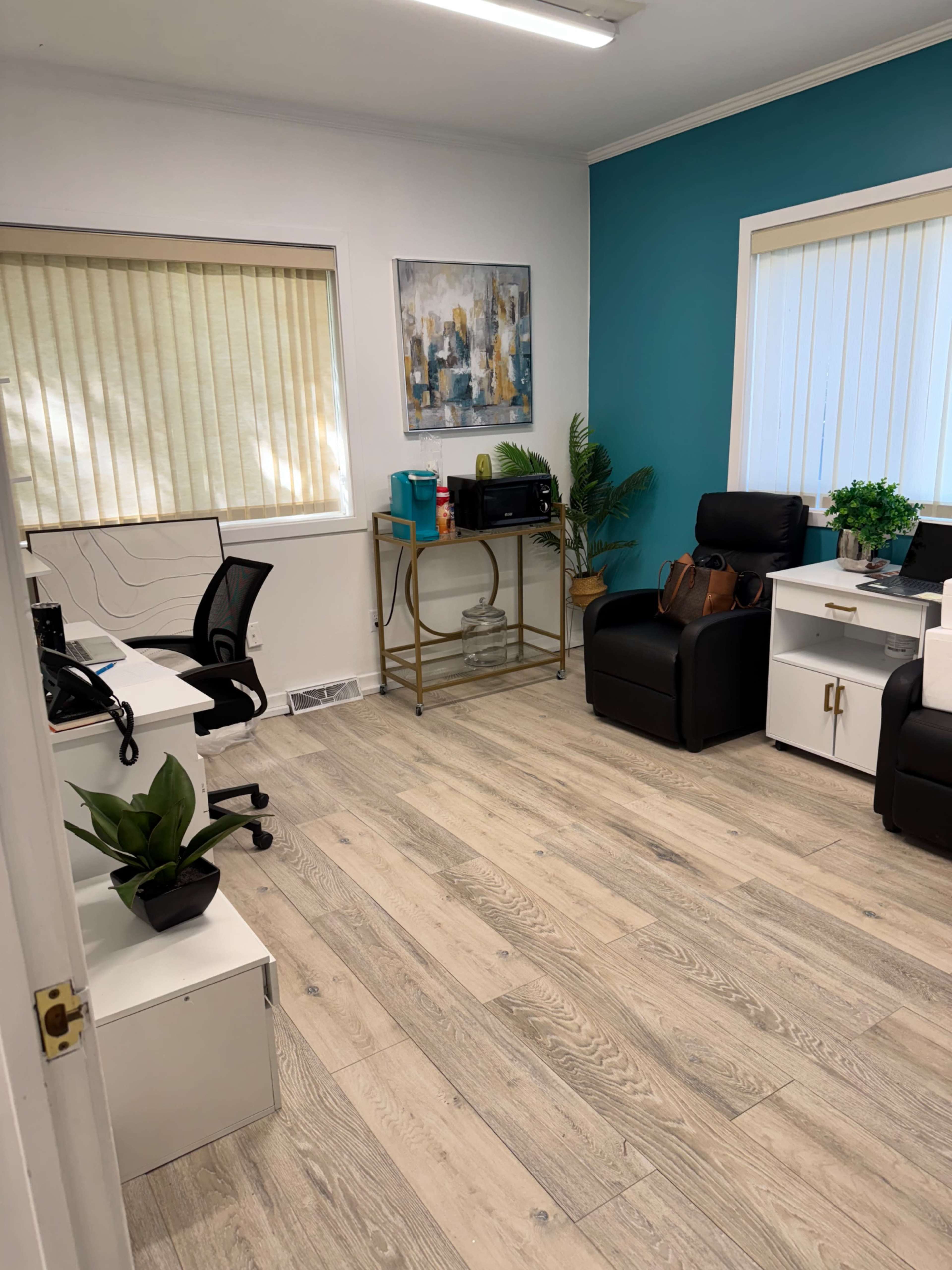 The image shows an office space with two workstations, a couch, a small table, and a decorative plant, featuring light wooden flooring and a blue accent wall.