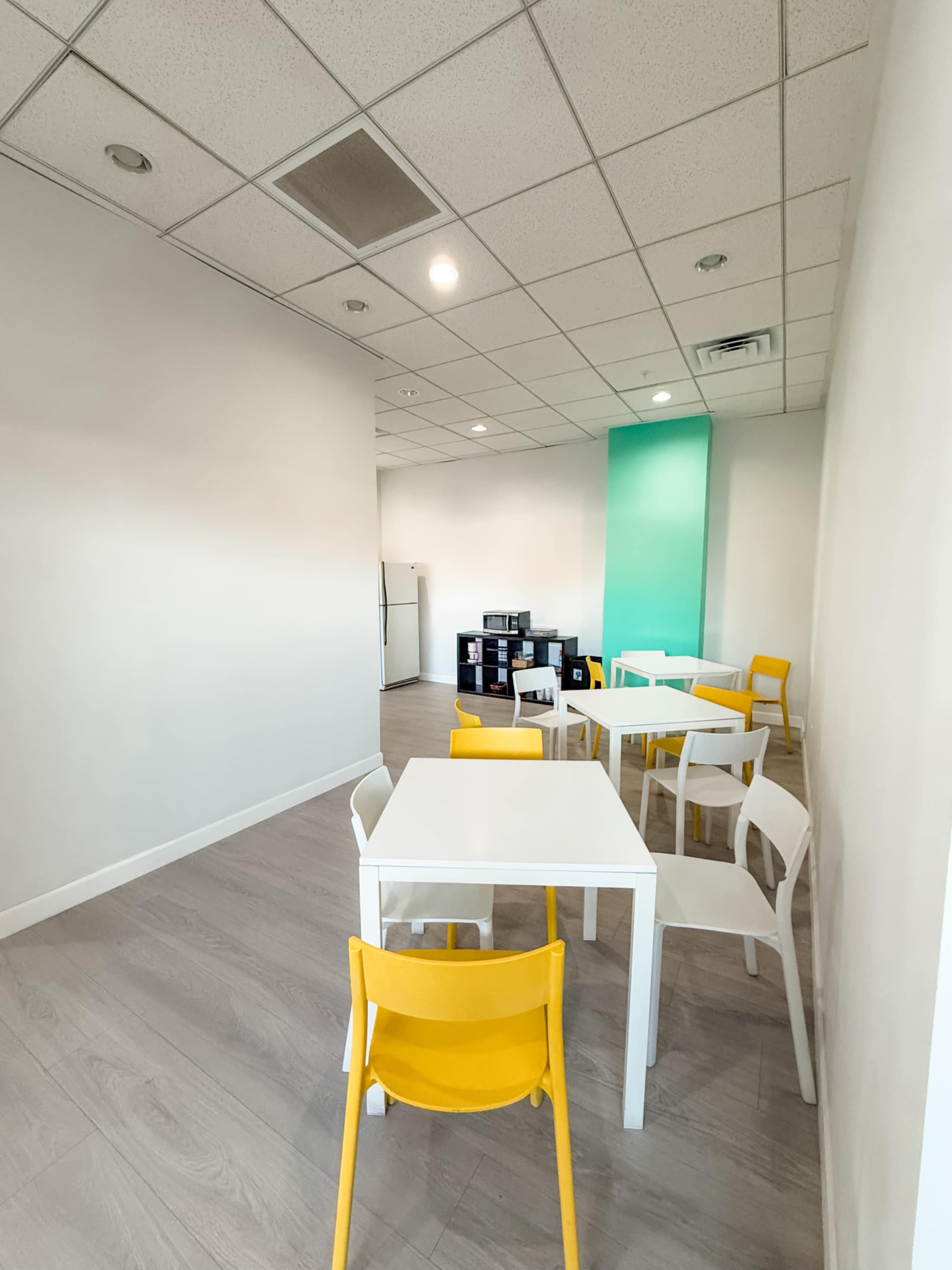 The image shows a brightly lit, minimalist room featuring several white tables and yellow chairs arranged around a light wooden floor.