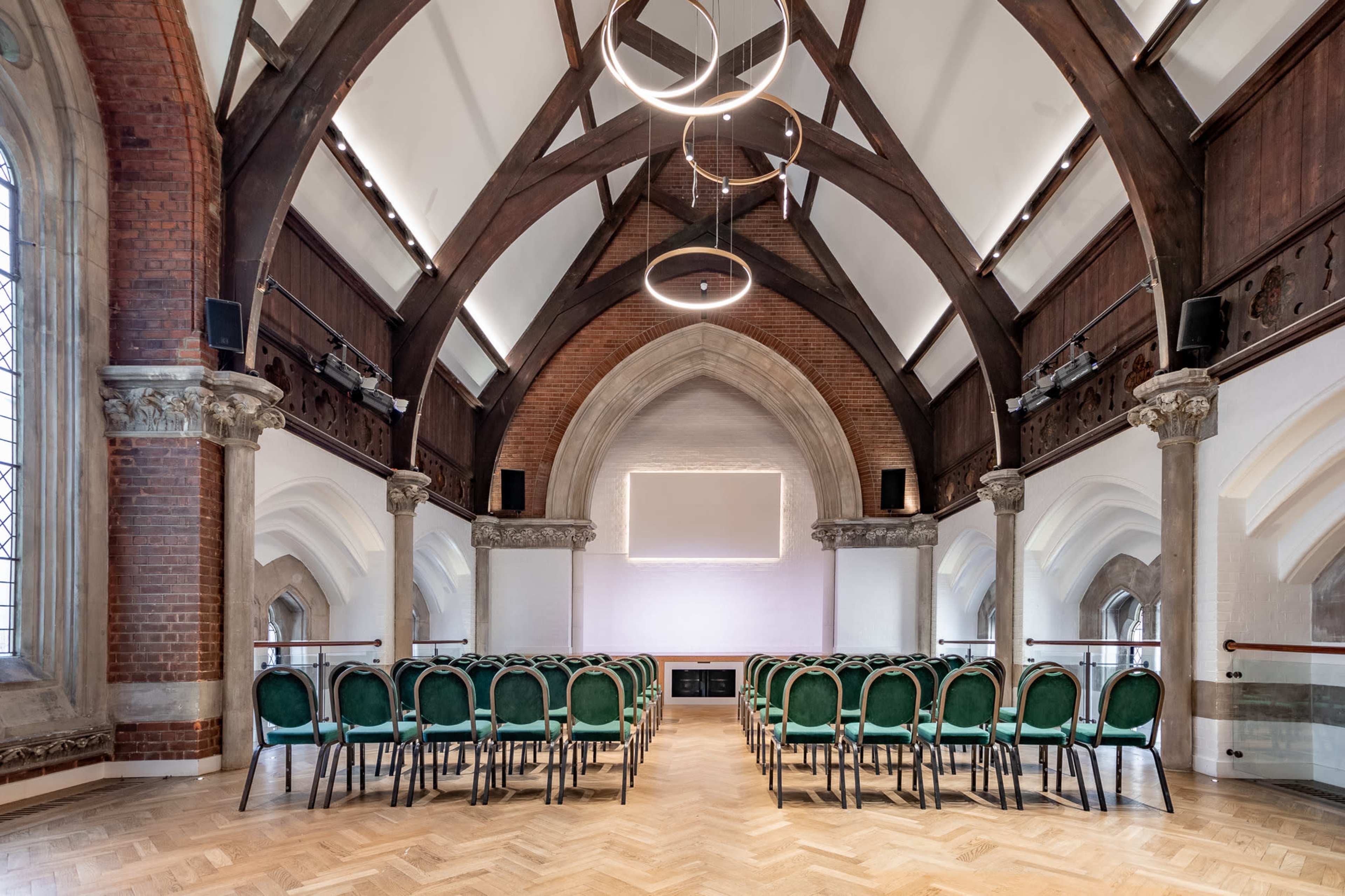 The image shows a spacious, elegantly designed hall with a sloped wooden ceiling, equipped with rows of green chairs arranged for an event.