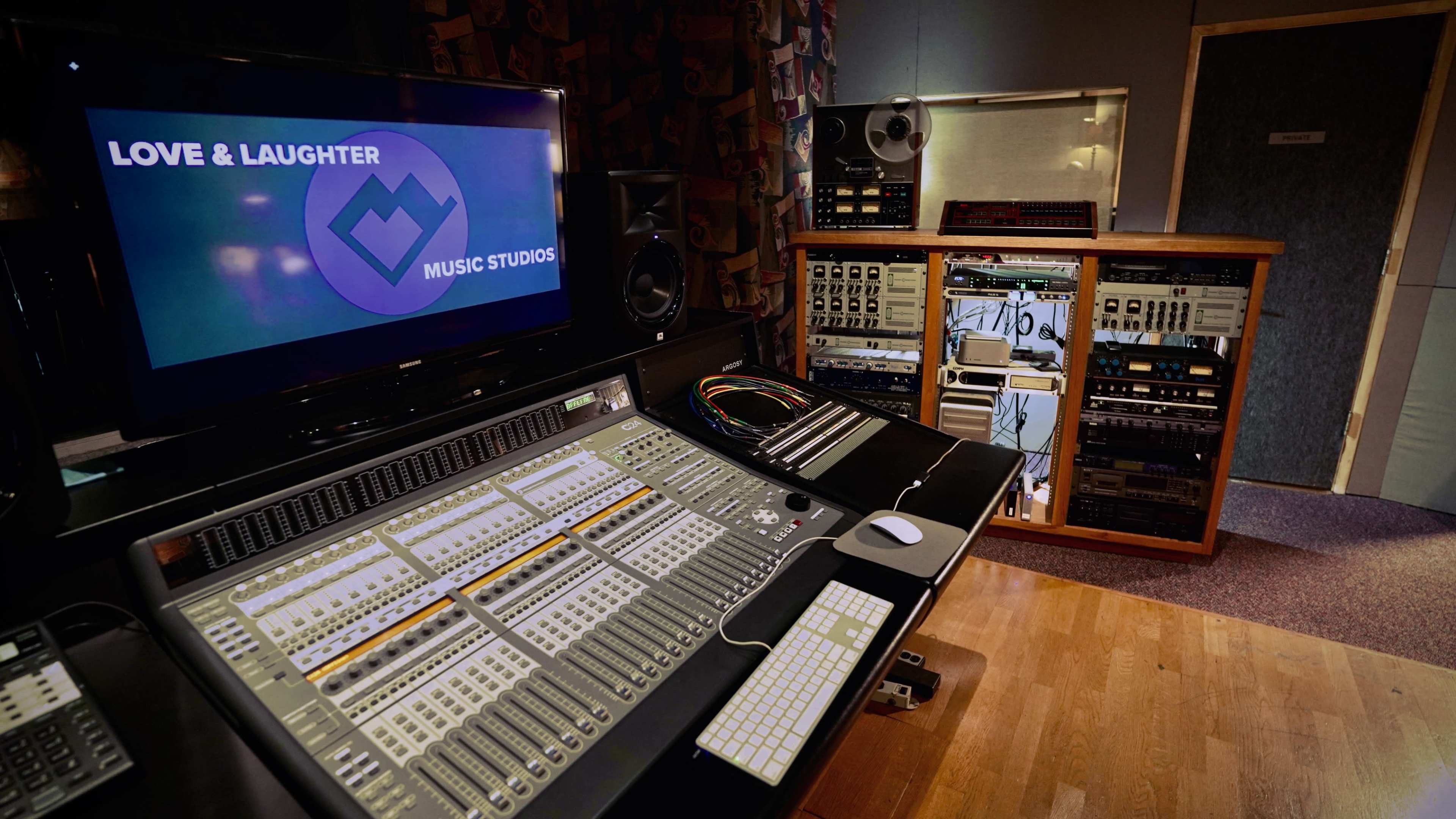 The image shows a music studio featuring a large mixing console with various control panels and equipment, alongside a computer screen displaying the studio's logo.