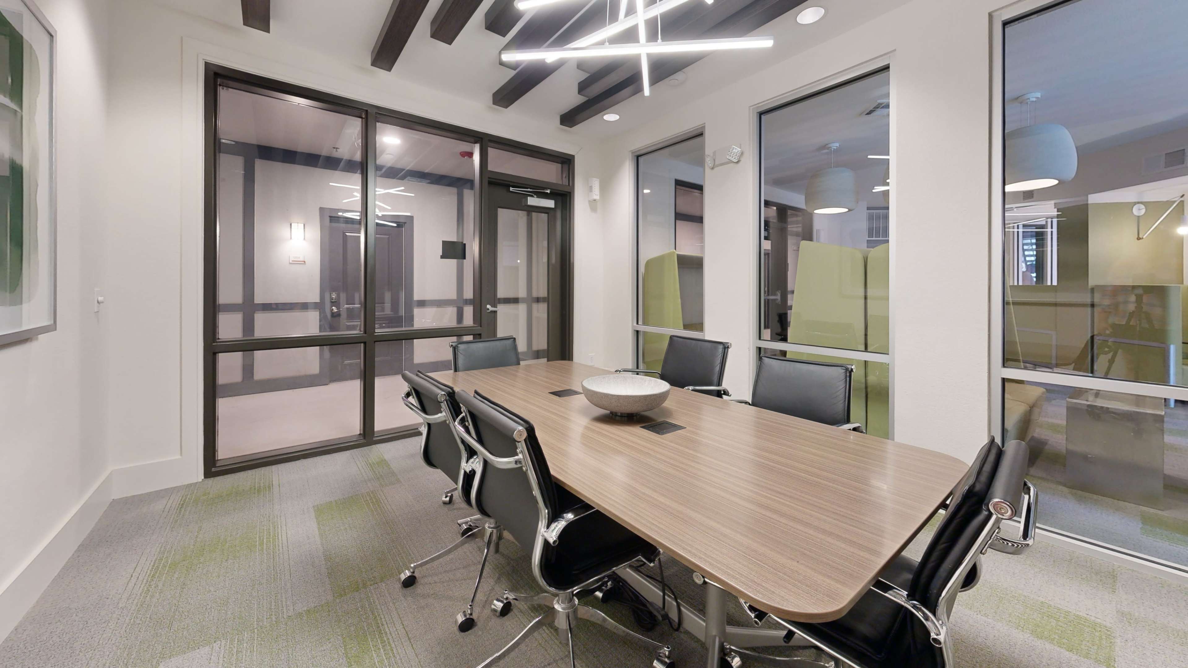 The image shows a modern conference room featuring a wooden table surrounded by black chairs, with glass walls providing visibility to an adjoining area.