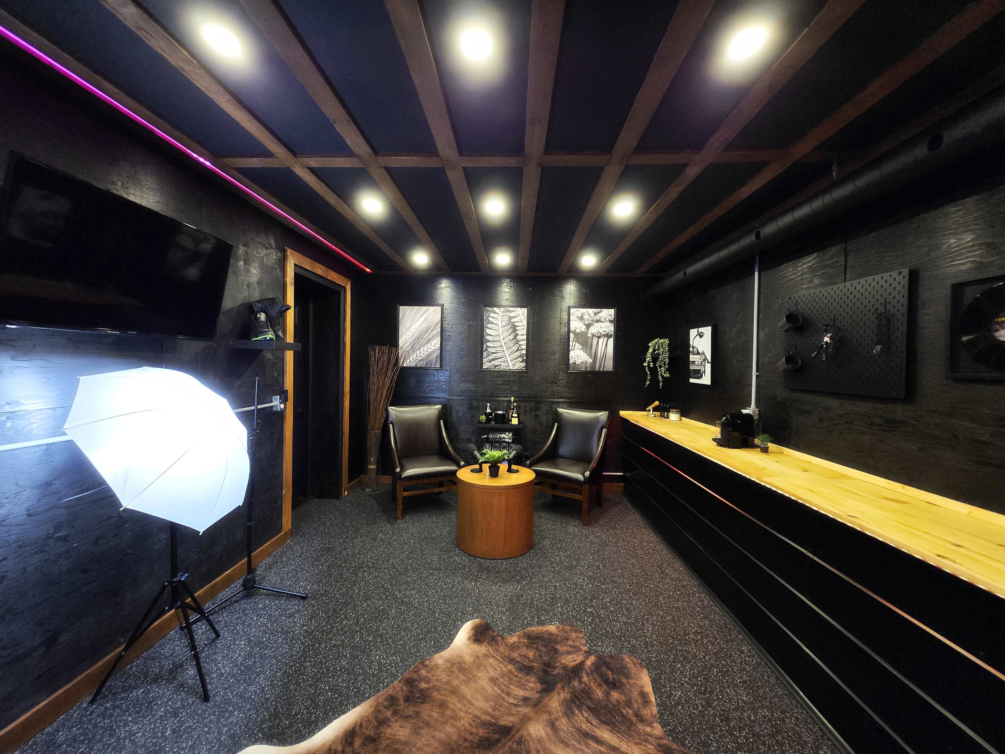 The image depicts a modern indoor lounge area featuring two chairs, a circular table, and various artistic wall decorations, along with professional lighting equipment.