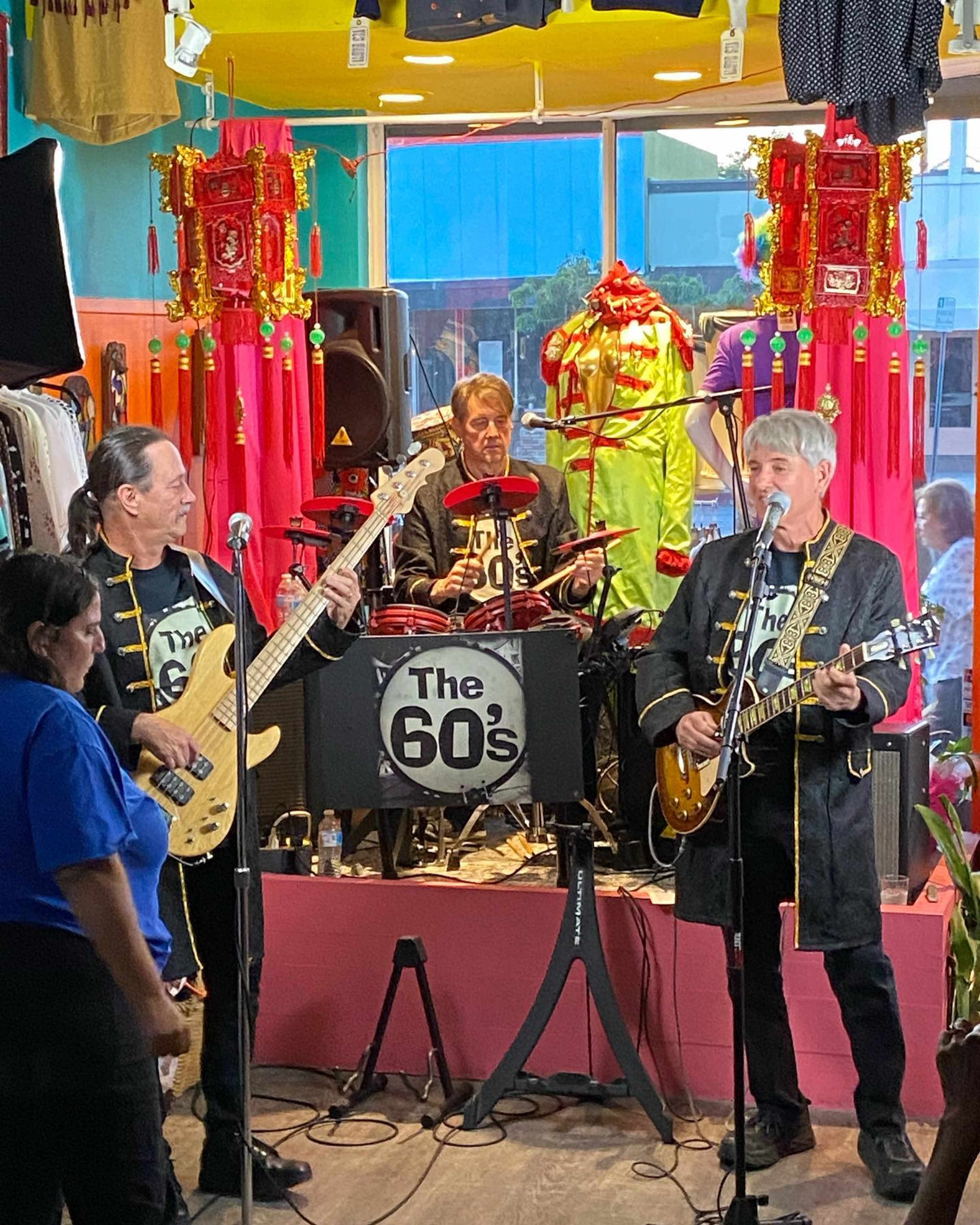 A band named "The 60's" performs in a colorful store setting, with two musicians on guitars and a drummer in the background.