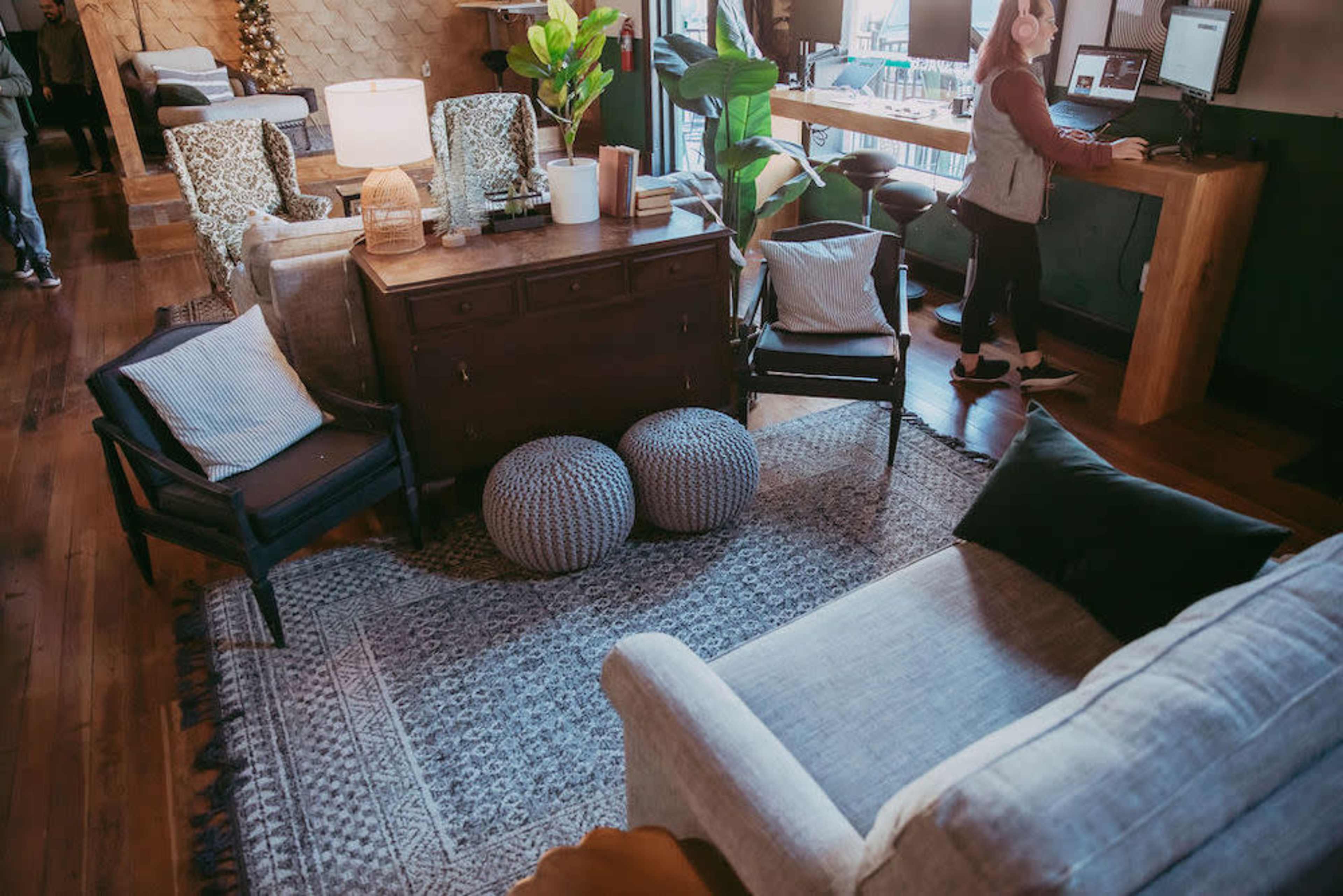The image shows a cozy interior with several seating areas, a wooden table, plants, and a person working at a desk.