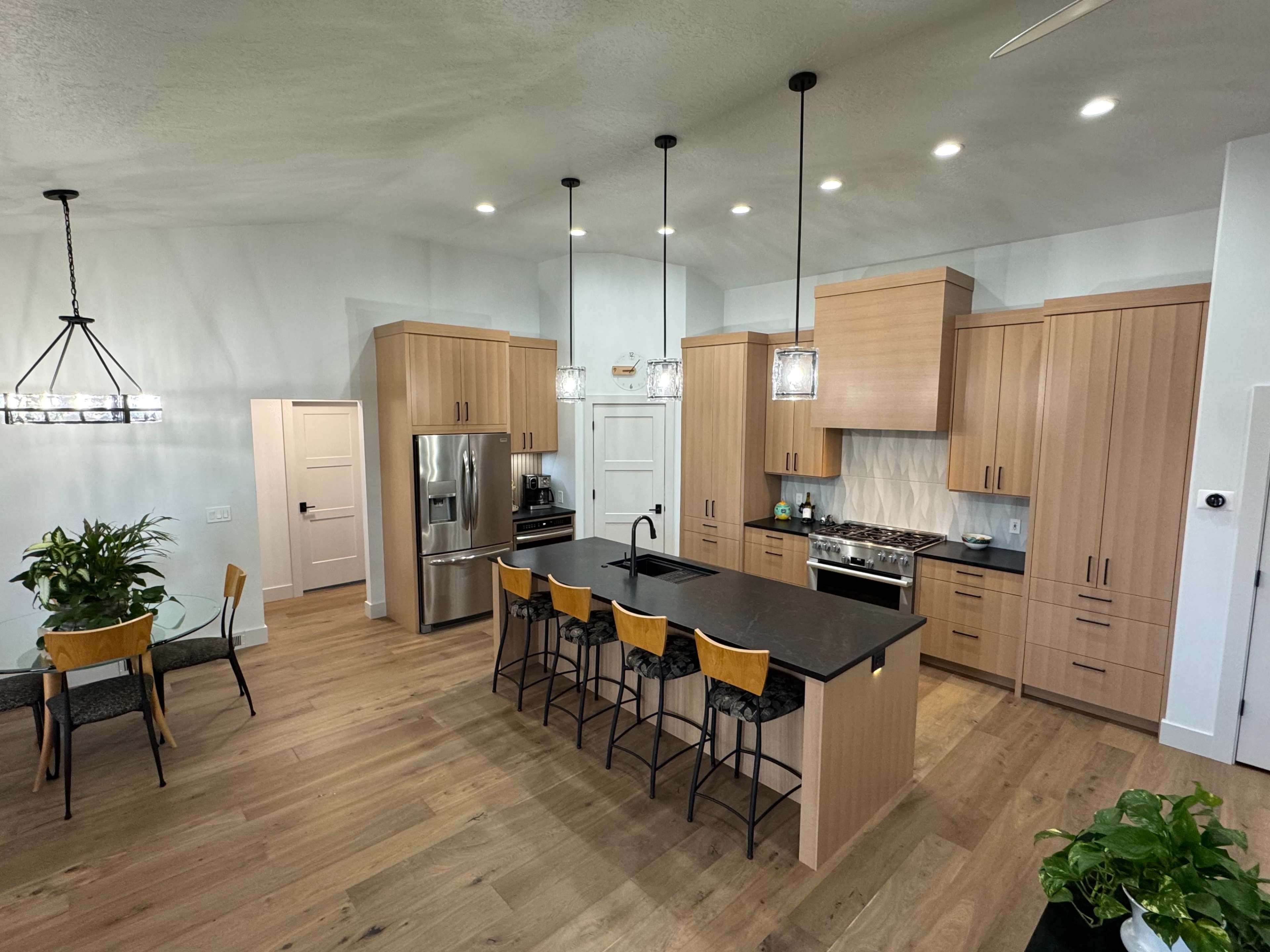 The image shows a modern kitchen with wooden cabinets, a large central island with seating, and a dining area featuring a round table and chairs.