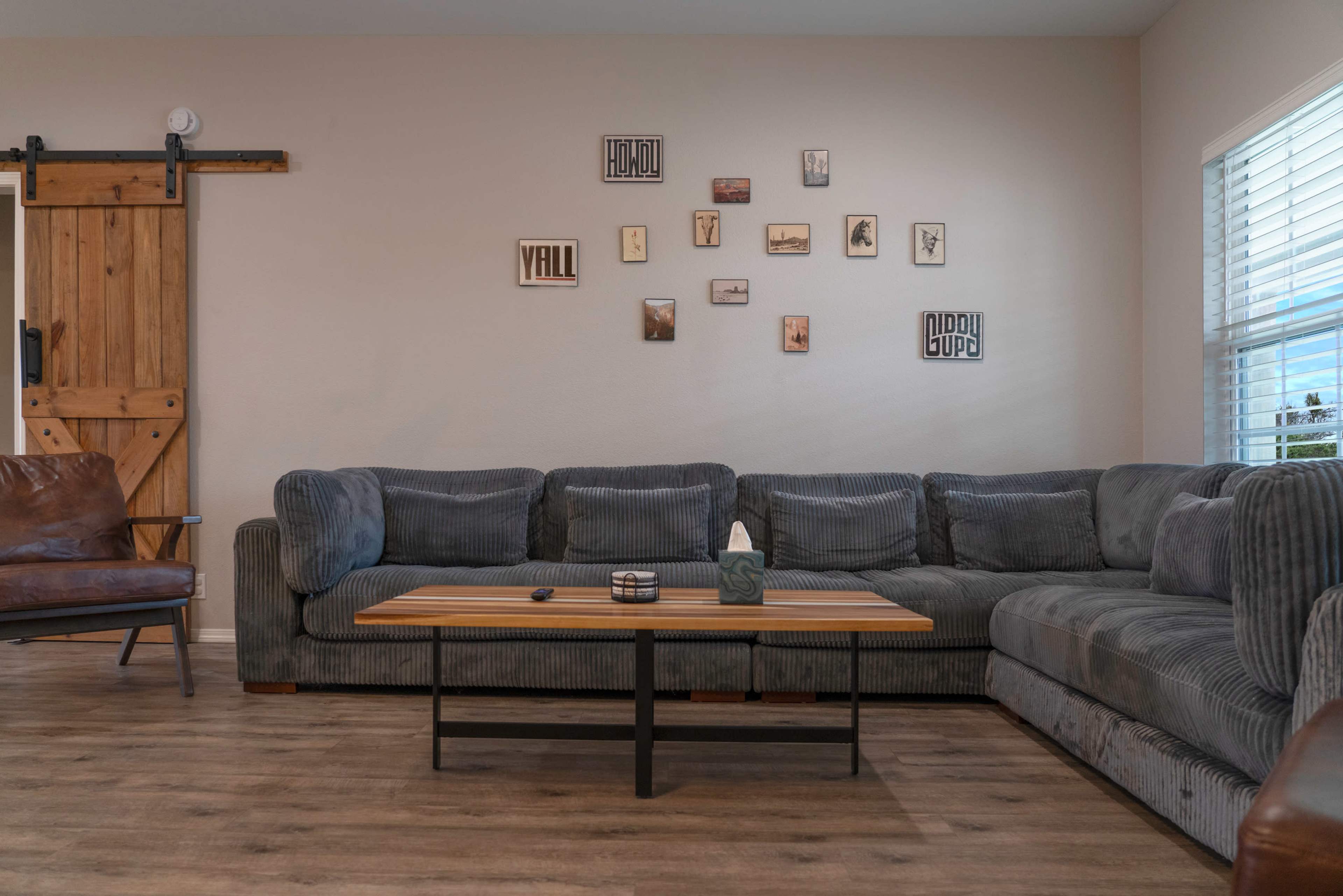 The image depicts a living room featuring a large sectional sofa, a coffee table, and a wall adorned with framed pictures, alongside a sliding barn door.
