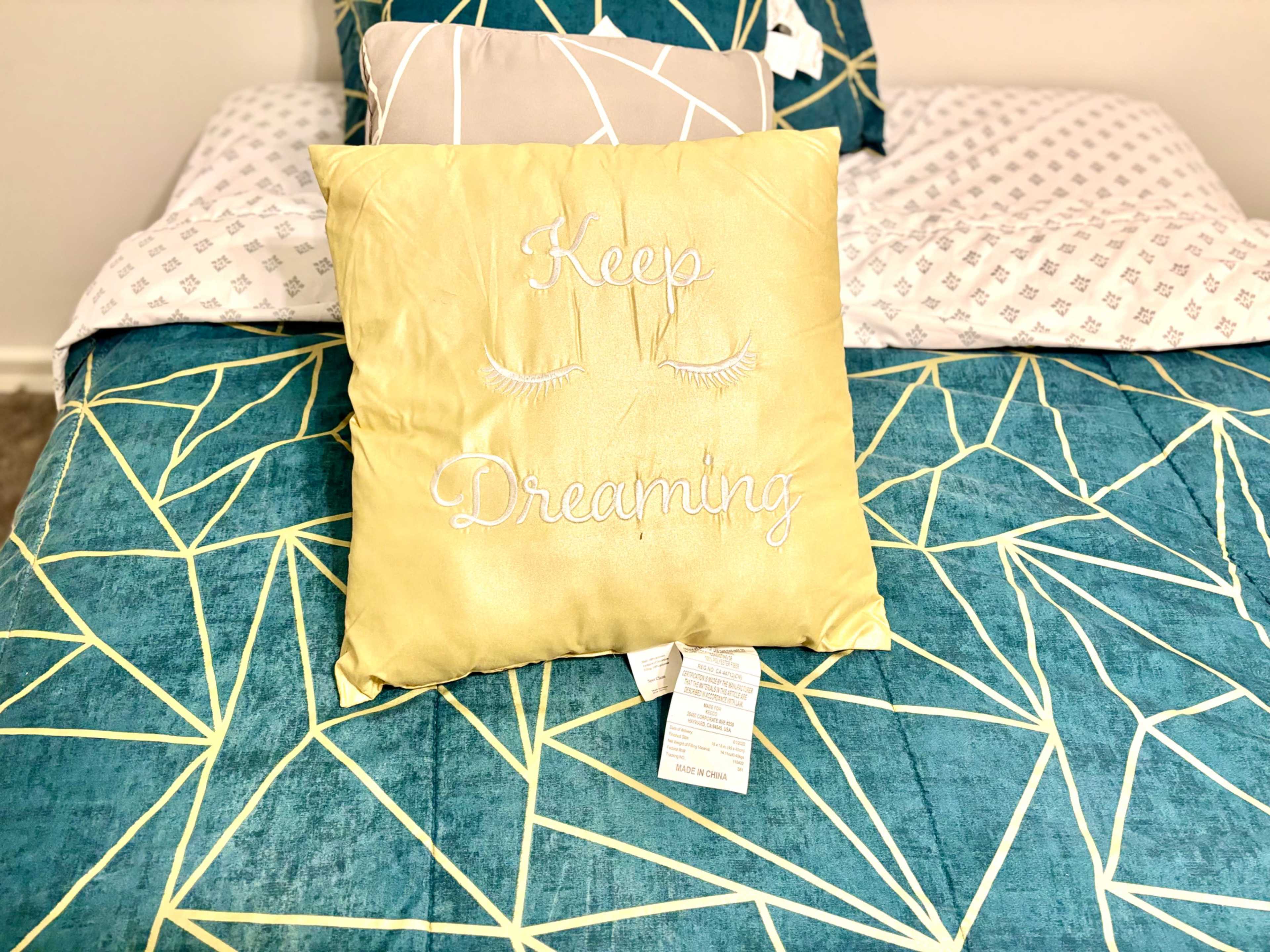 A yellow pillow with the text "Keep Dreaming" is placed on a patterned turquoise bedspread.