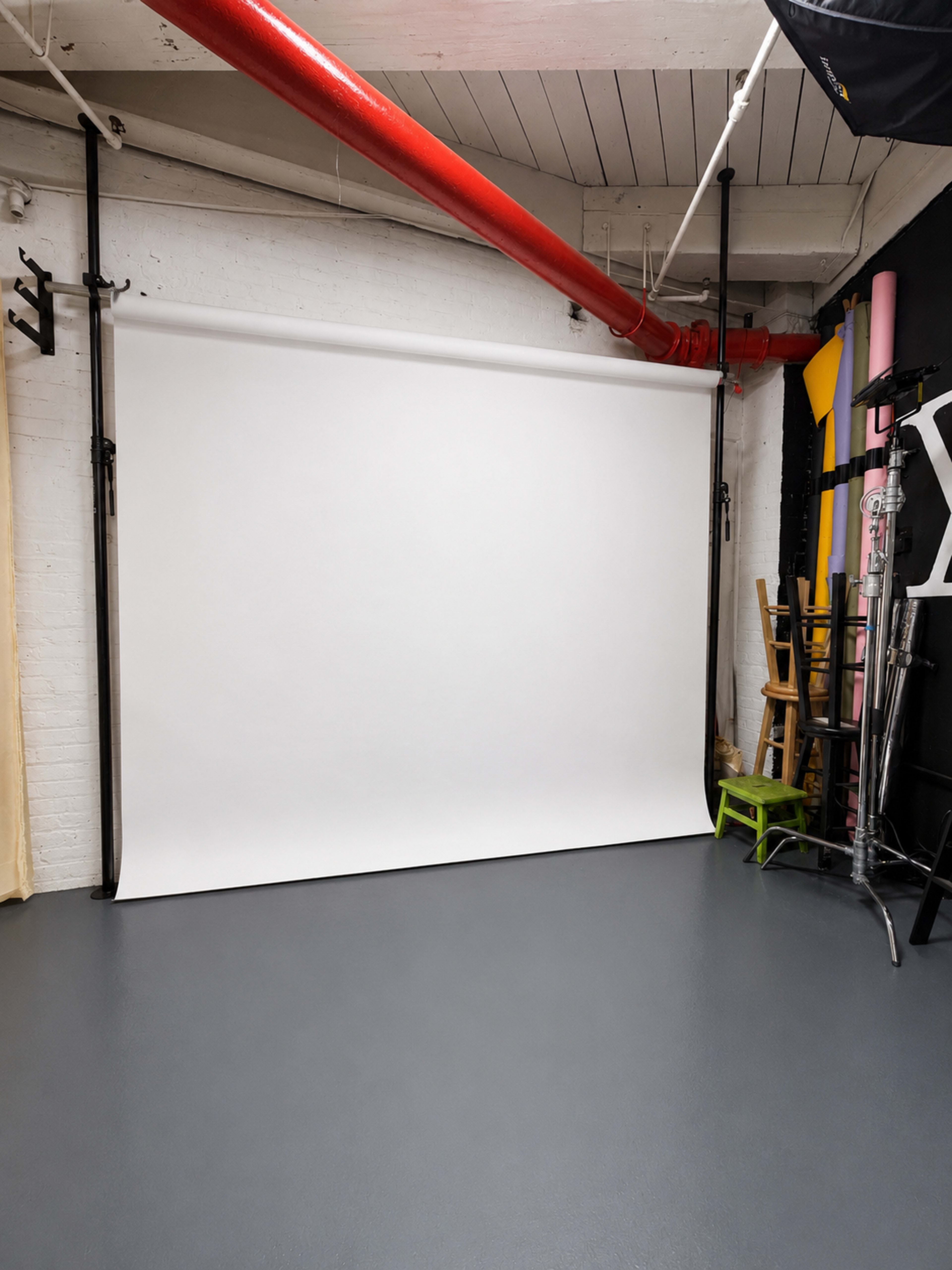 The image shows a photography studio with a white backdrop, gray floor, and various equipment, including chairs and a tall ladder, stored against the walls.