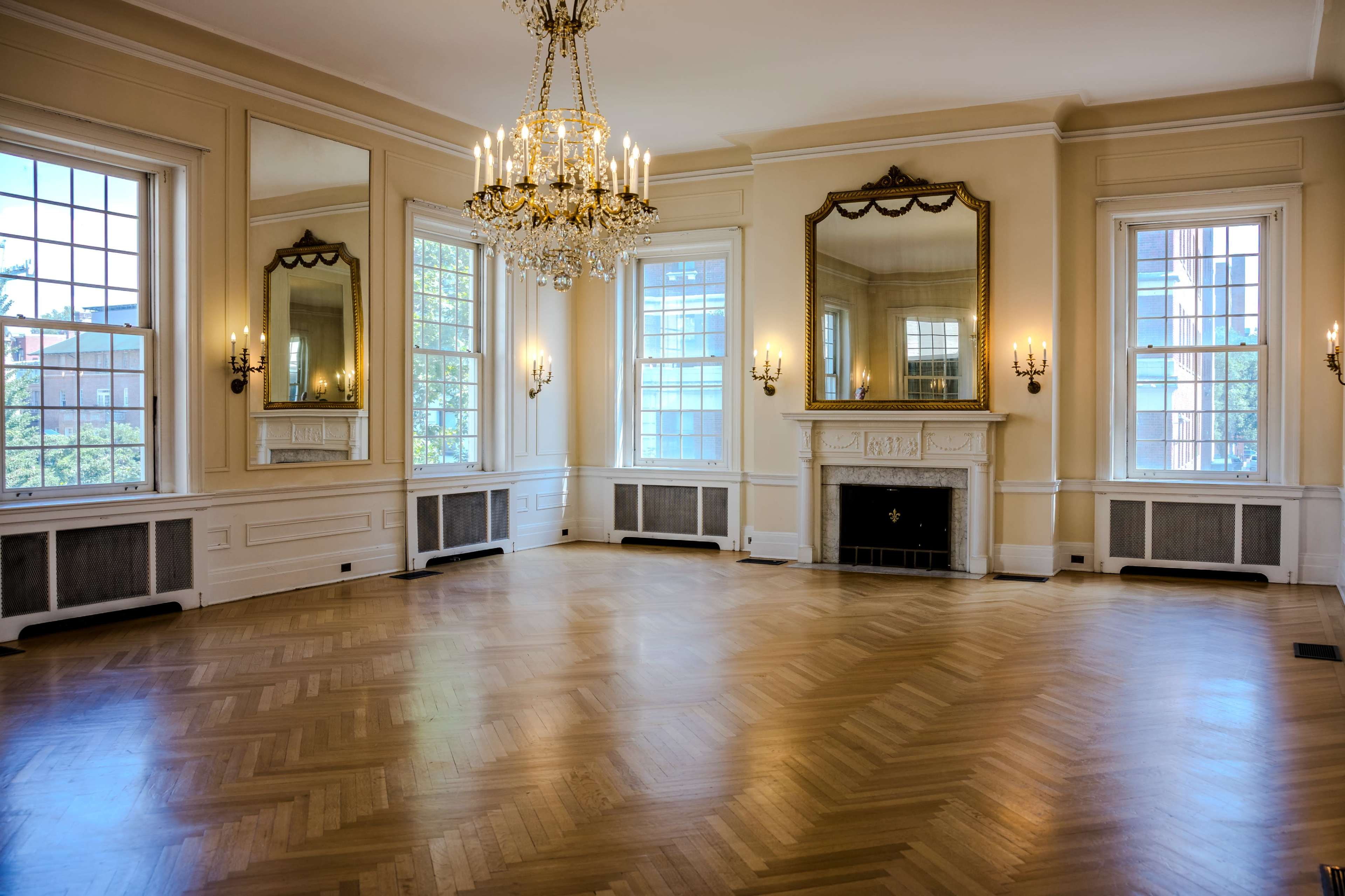 The room features wooden herringbone flooring, large windows, ornate wall mirrors, a chandelier, and a decorative fireplace.
