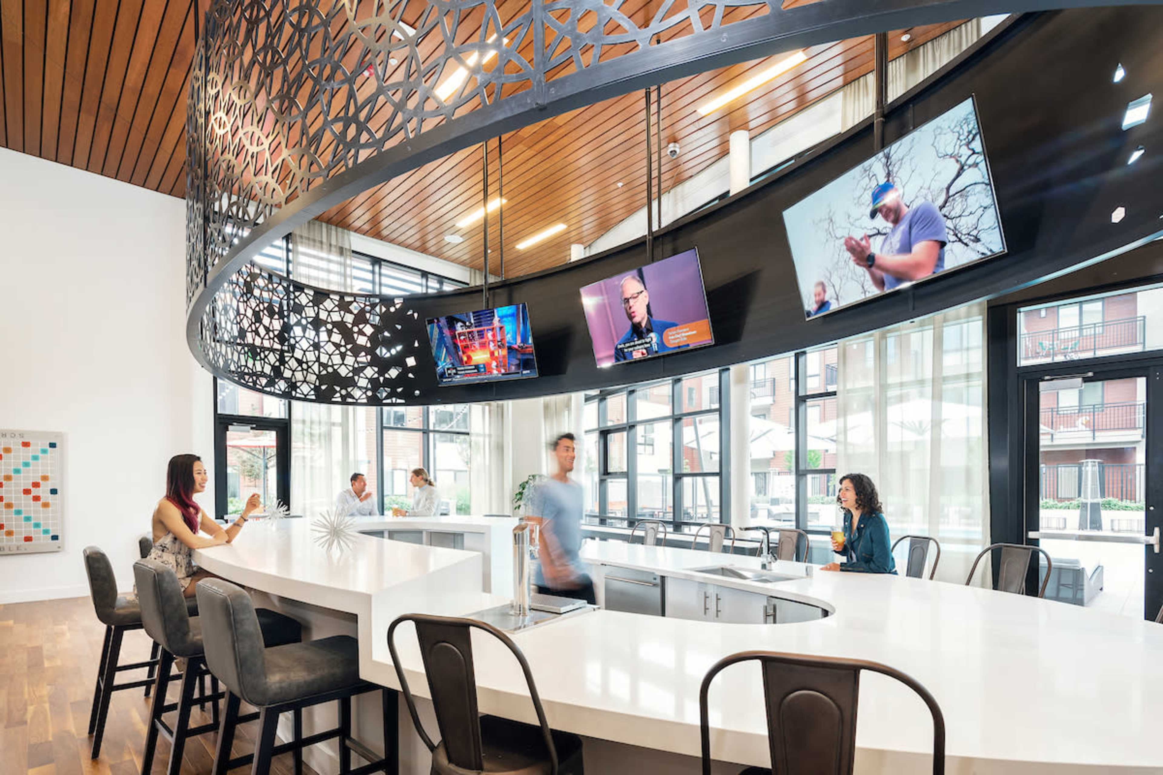 A modern café interior features a white countertop bar with high stools, large windows letting in natural light, and multiple flat-screen TVs mounted on the wall.