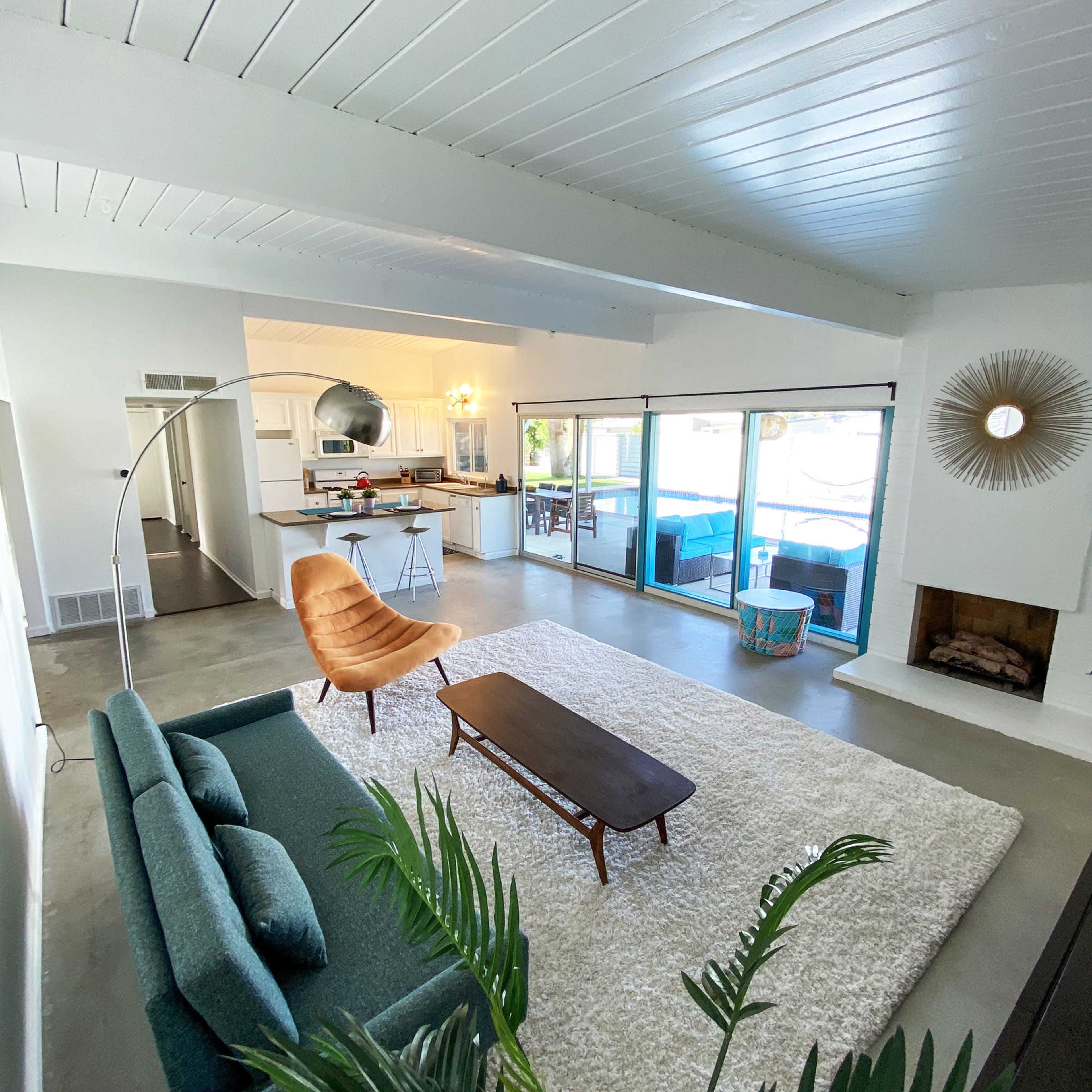 A spacious living area features a green sofa, a wooden coffee table, and a large rug, with a kitchen visible in the background and large sliding glass doors leading outside.