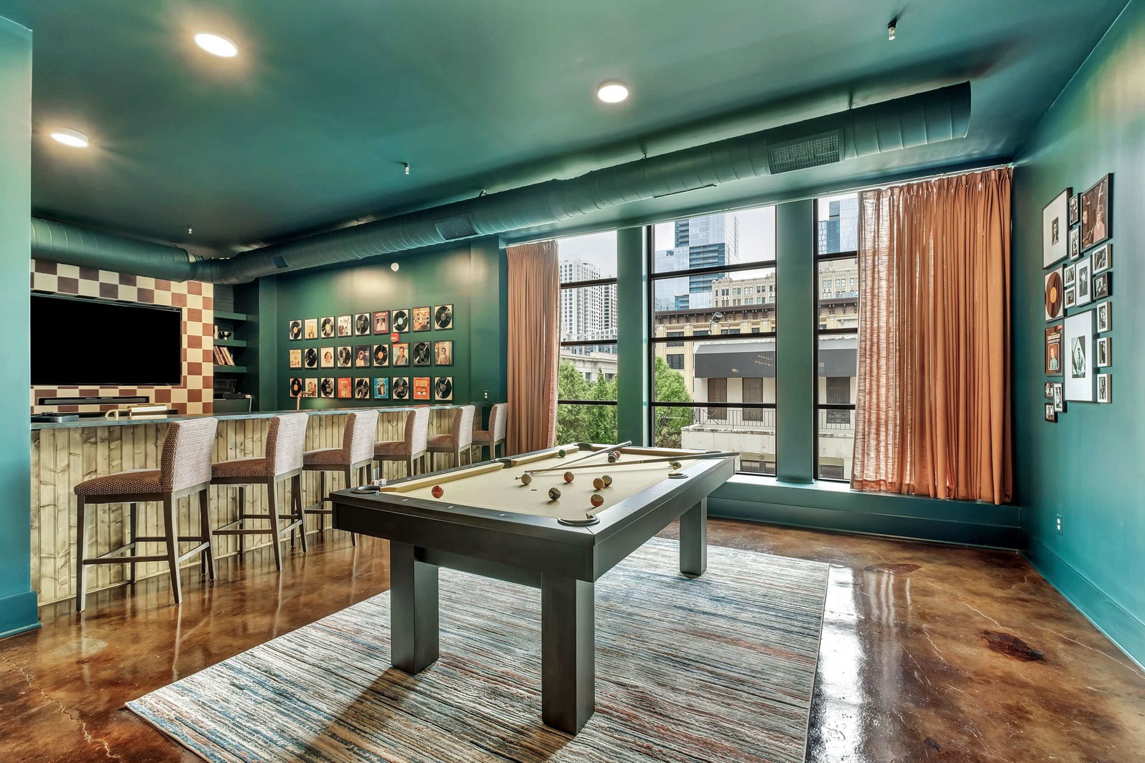 The image shows a recreational room featuring a billiards table, a bar area with seating, and large windows that overlook a cityscape.