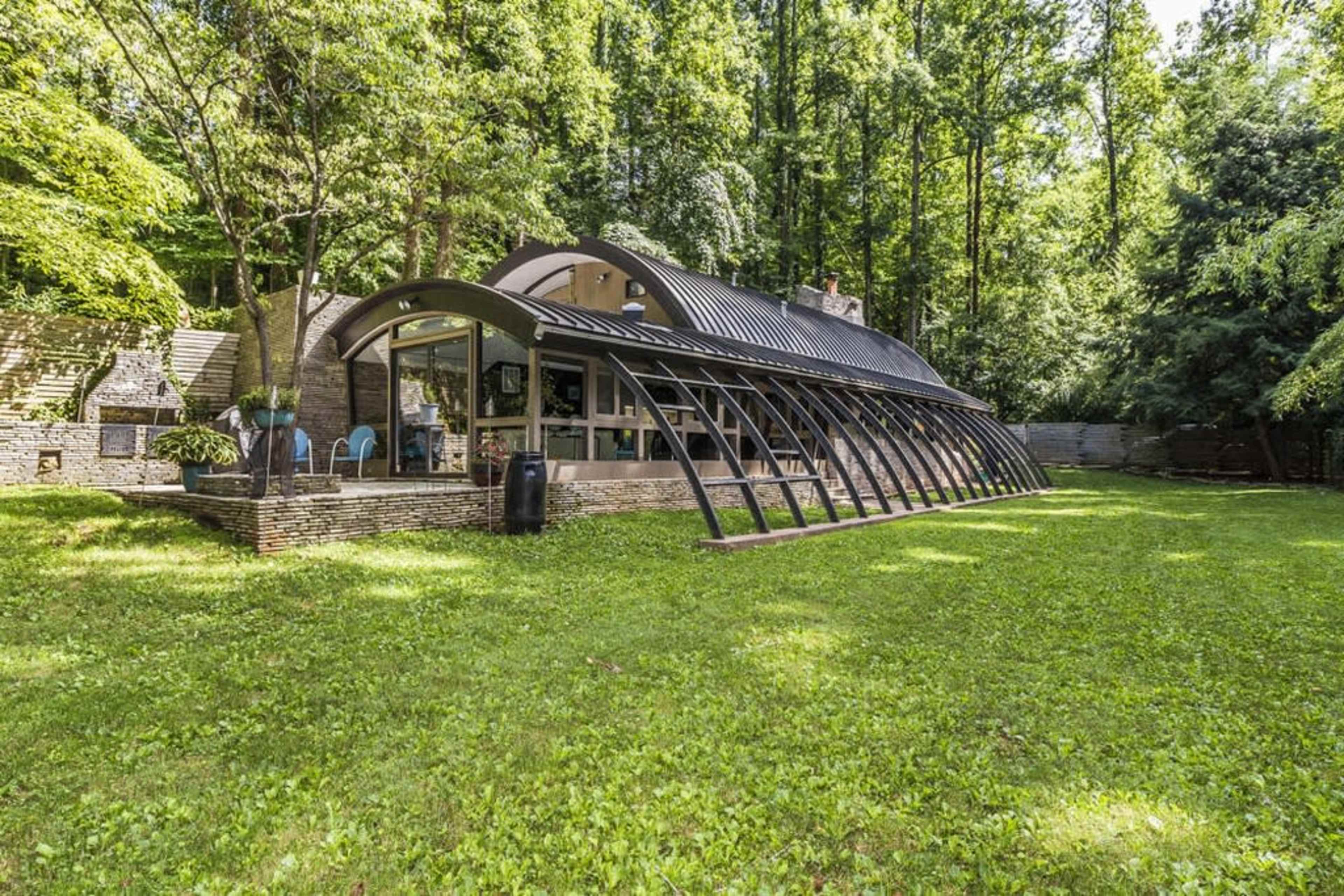 A modern, curved glass and metal structure is set in a lush green yard surrounded by trees.
