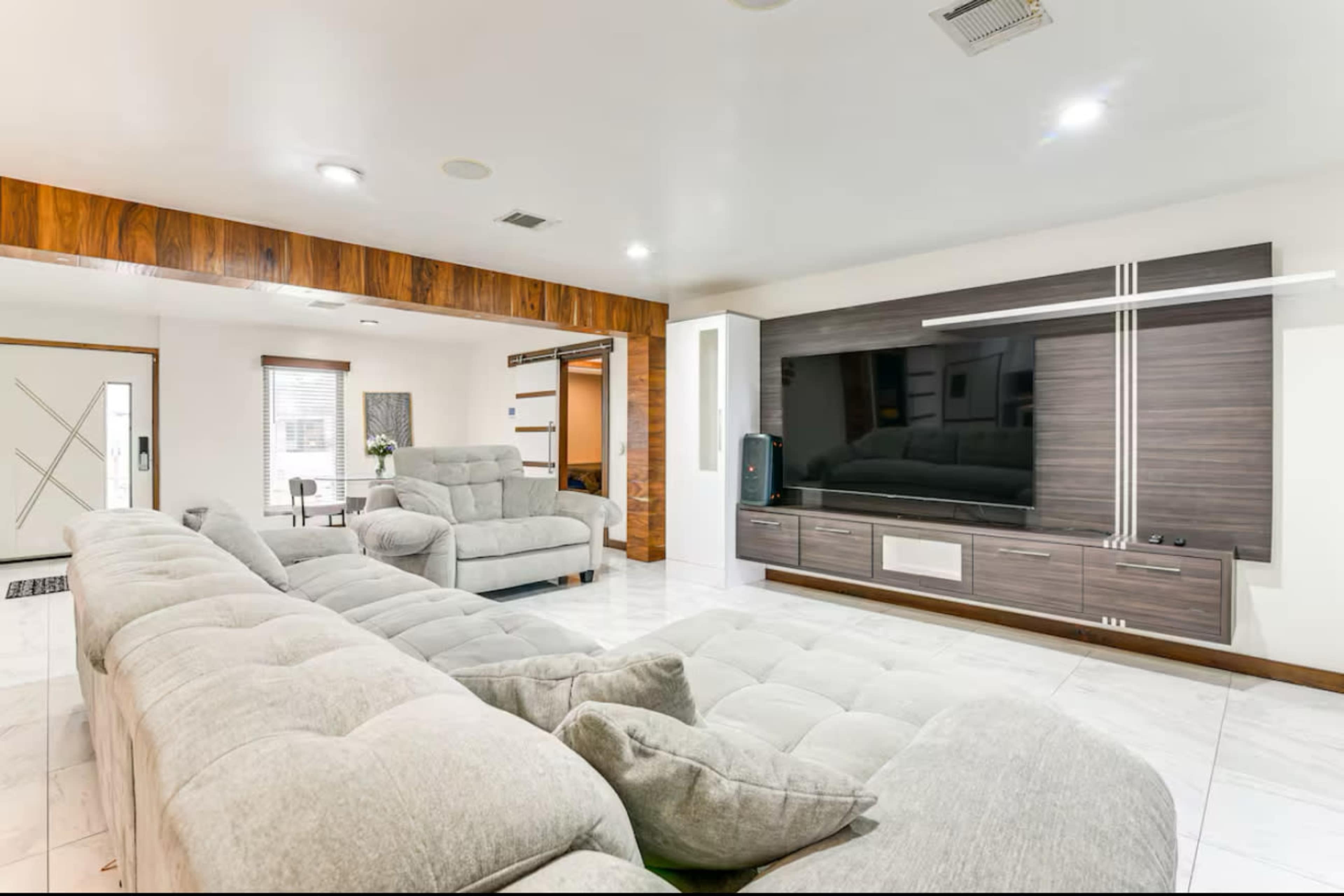 The image shows a modern living room with a large, gray sectional sofa facing a wall-mounted television and wooden cabinetry.