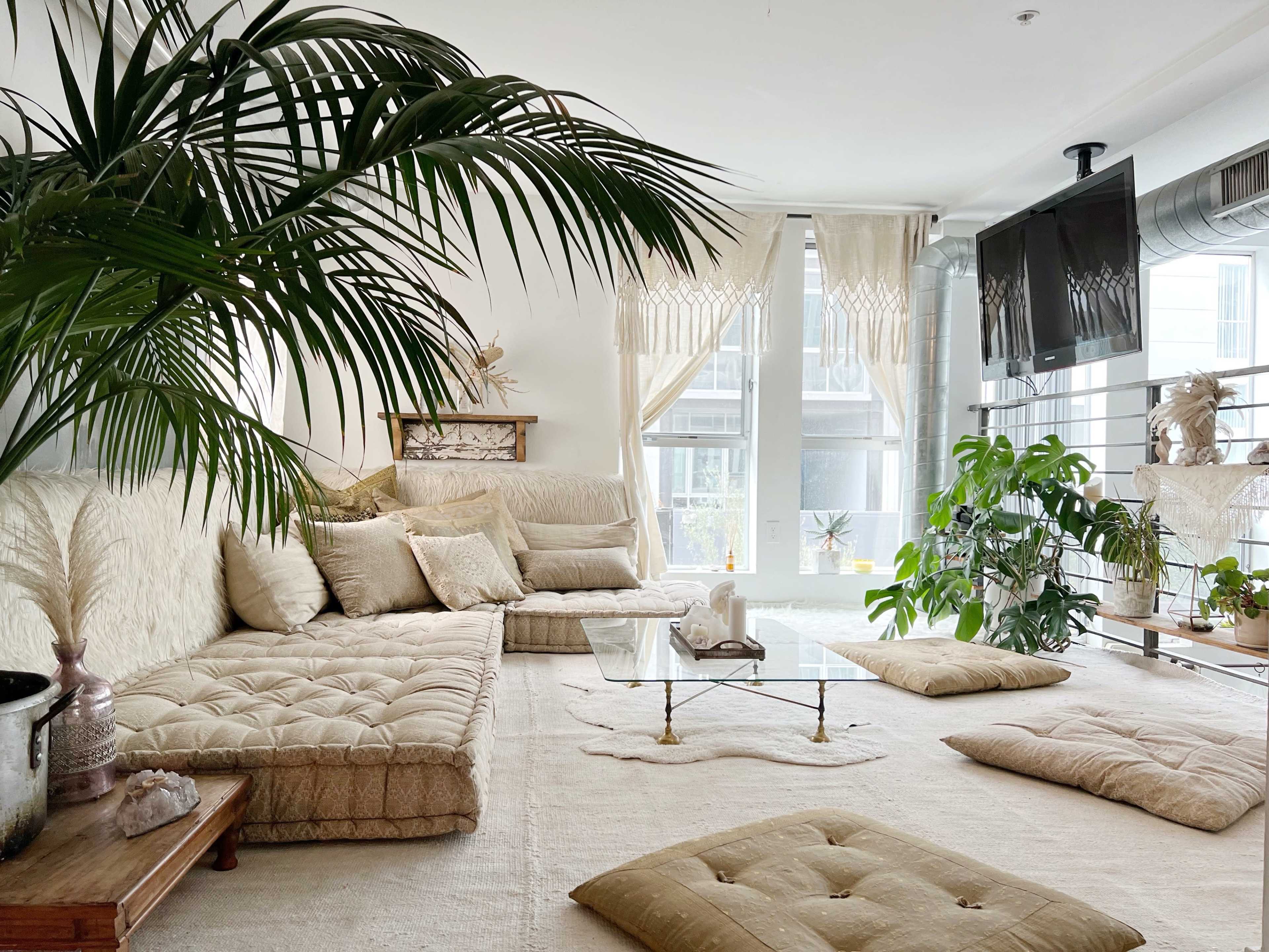 The room features a spacious, light-filled living area with a large, plush sectional sofa, several floor cushions, a glass coffee table, and an abundance of indoor plants.