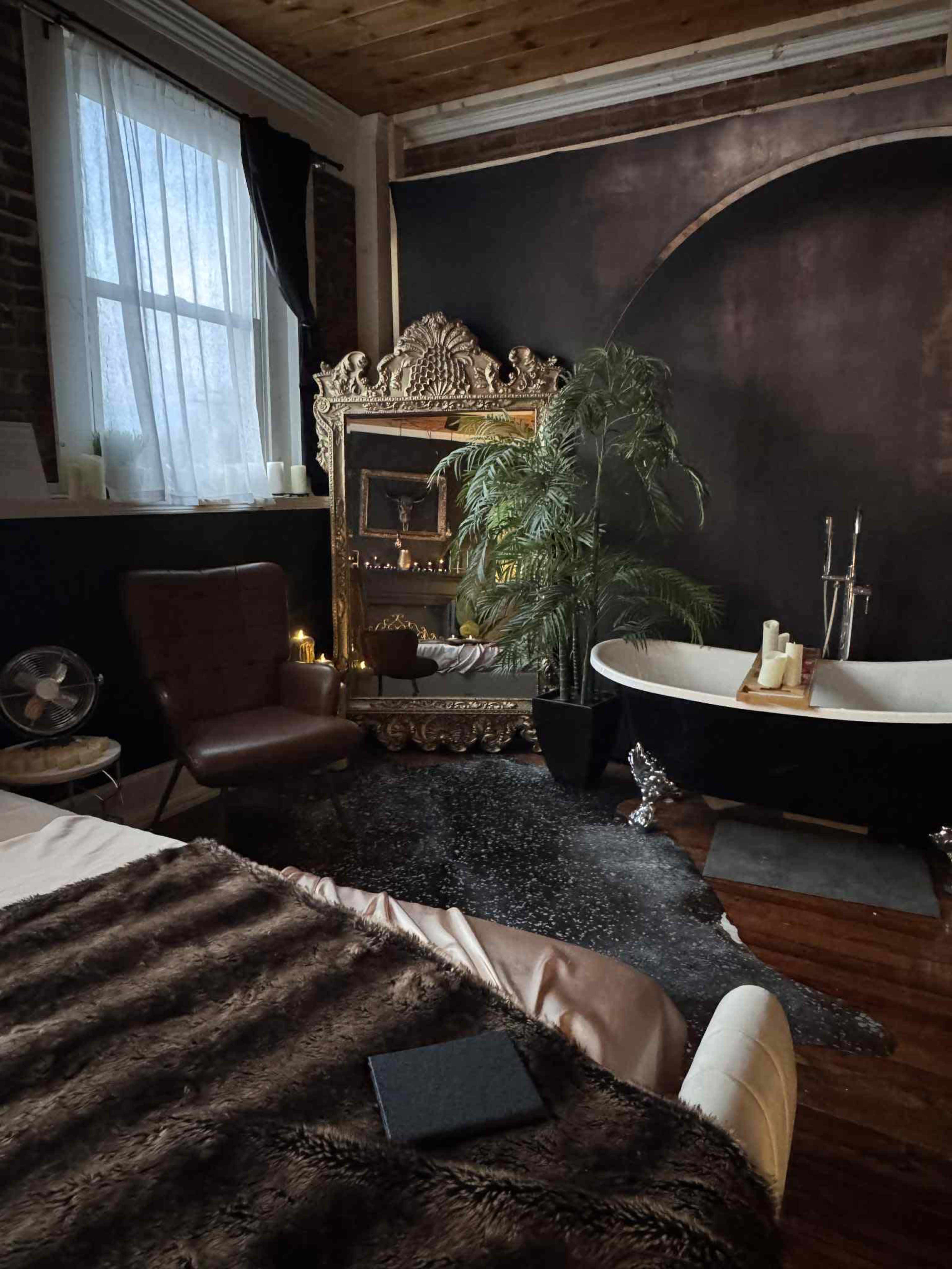 A cozy bedroom features a large, ornate mirror, a vintage armchair, a bathtub with candles, and a lush plant, all set against dark walls and wooden floors.