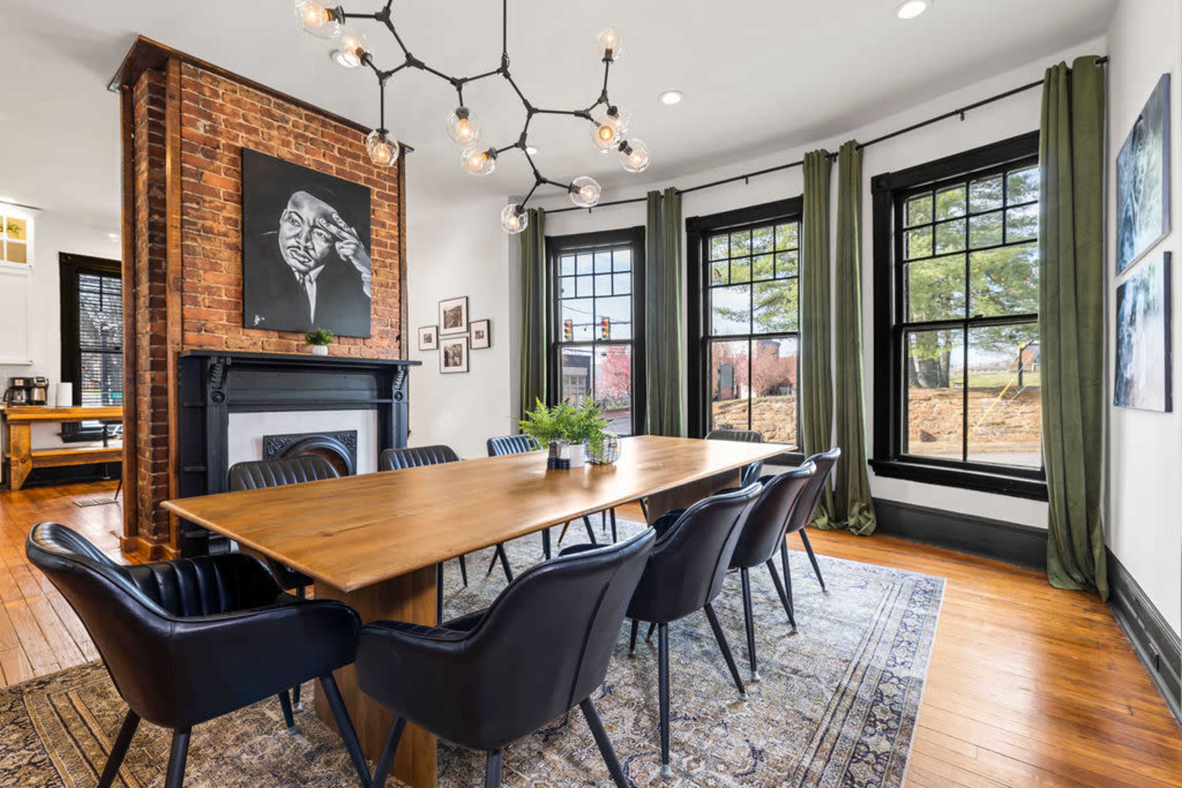 A dining room featuring a long wooden table surrounded by black chairs, with a large brick fireplace, artwork on the wall, and large windows dressed in green curtains.