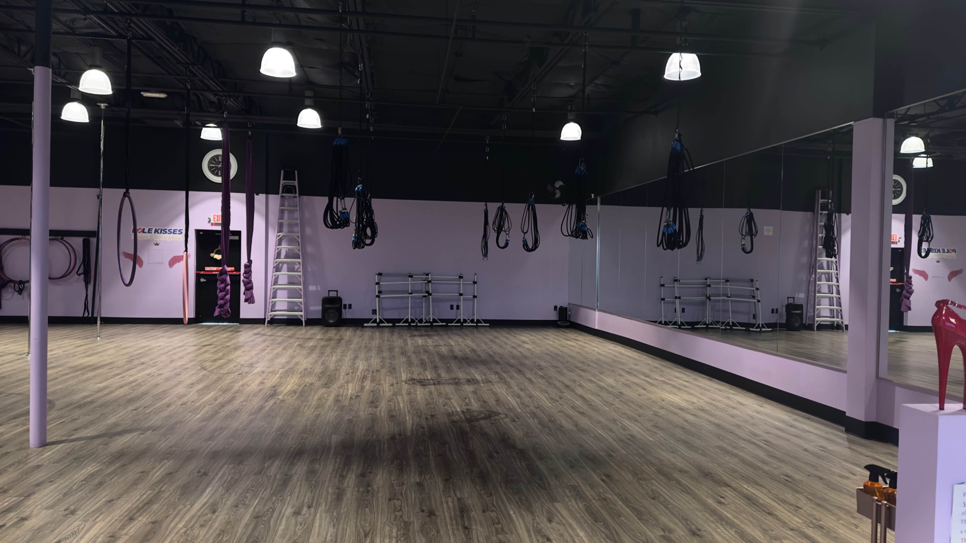 A fitness studio with a spacious wooden floor, mirrors on one wall, and various straps hanging from the ceiling.