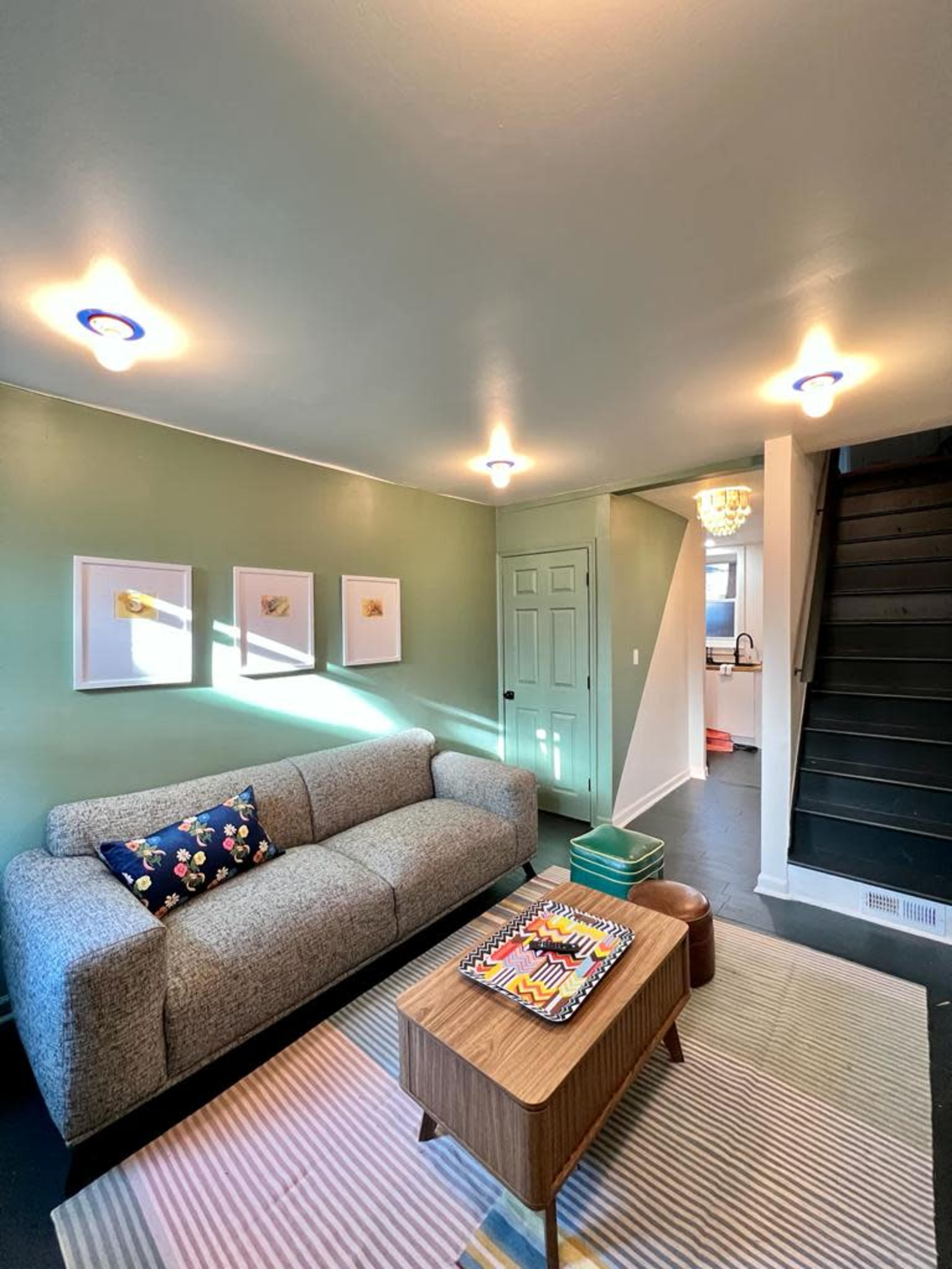 A living room with a gray sofa, a wooden coffee table, and green walls, featuring framed artwork and a staircase in the background.