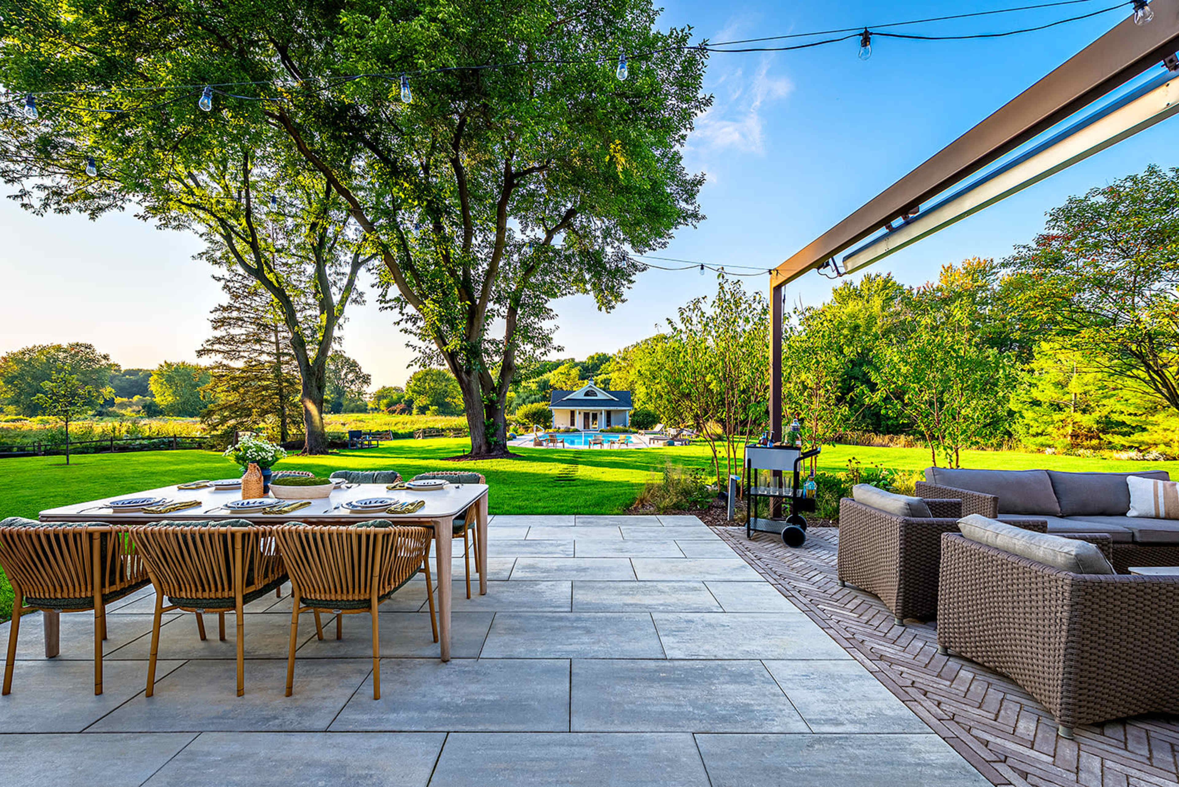 A patio features a dining table set for outdoor meals, with trees and a pool visible in the background.