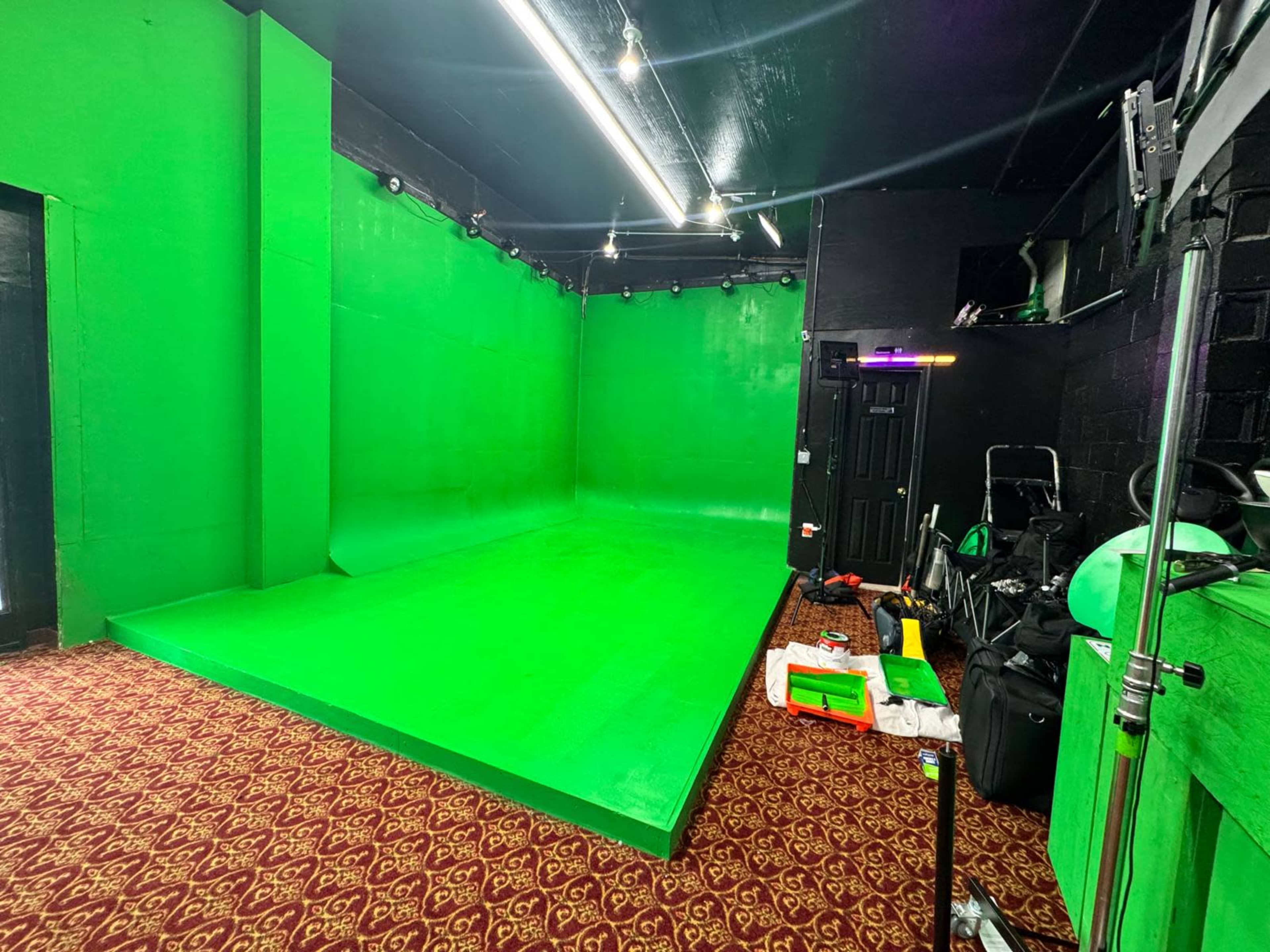 The image shows a green screen studio with a textured carpet, lighting fixtures, and various equipment scattered on the floor.