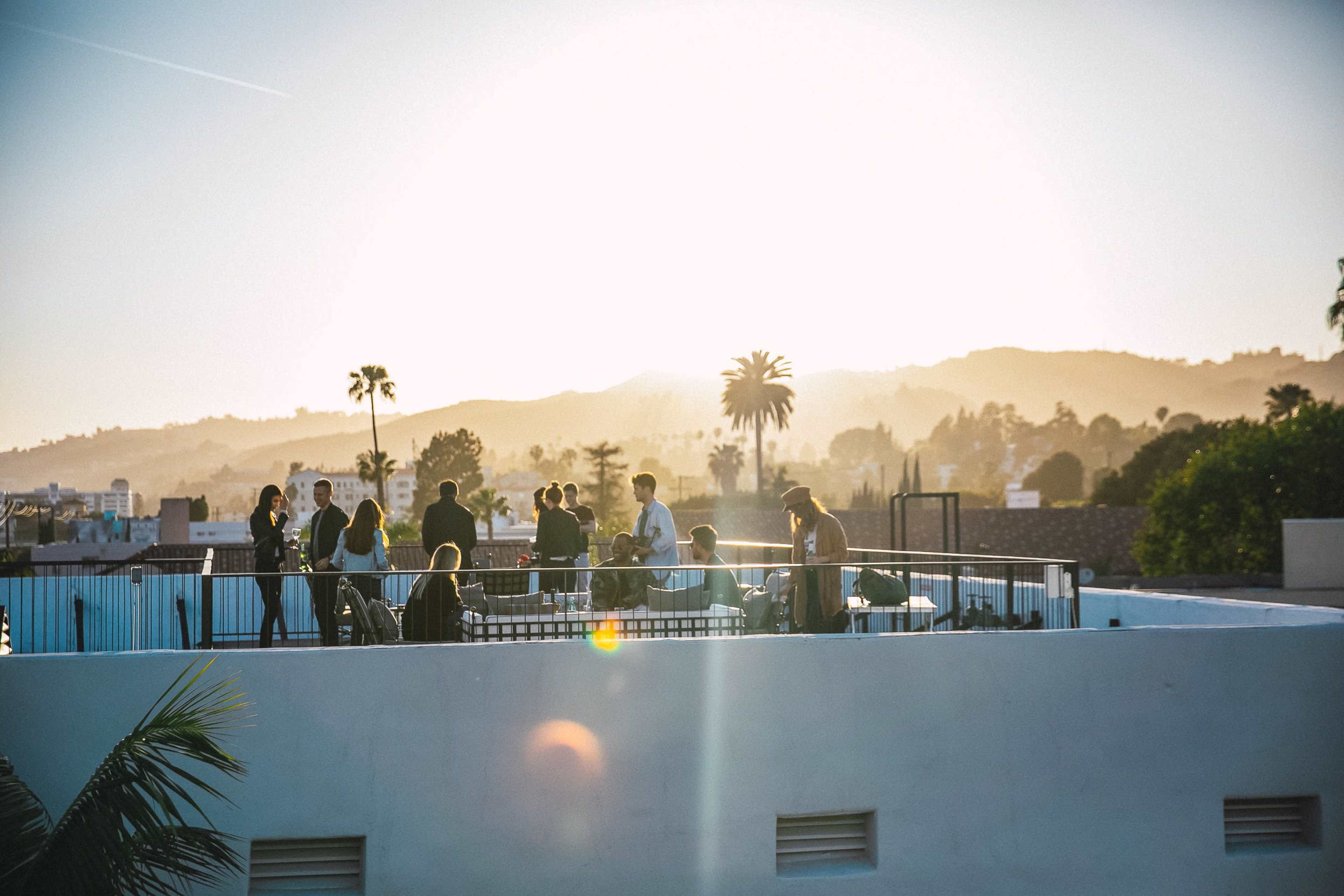 A group of people gathers on a rooftop, silhouetted against a sunset backdrop of hills and palm trees.