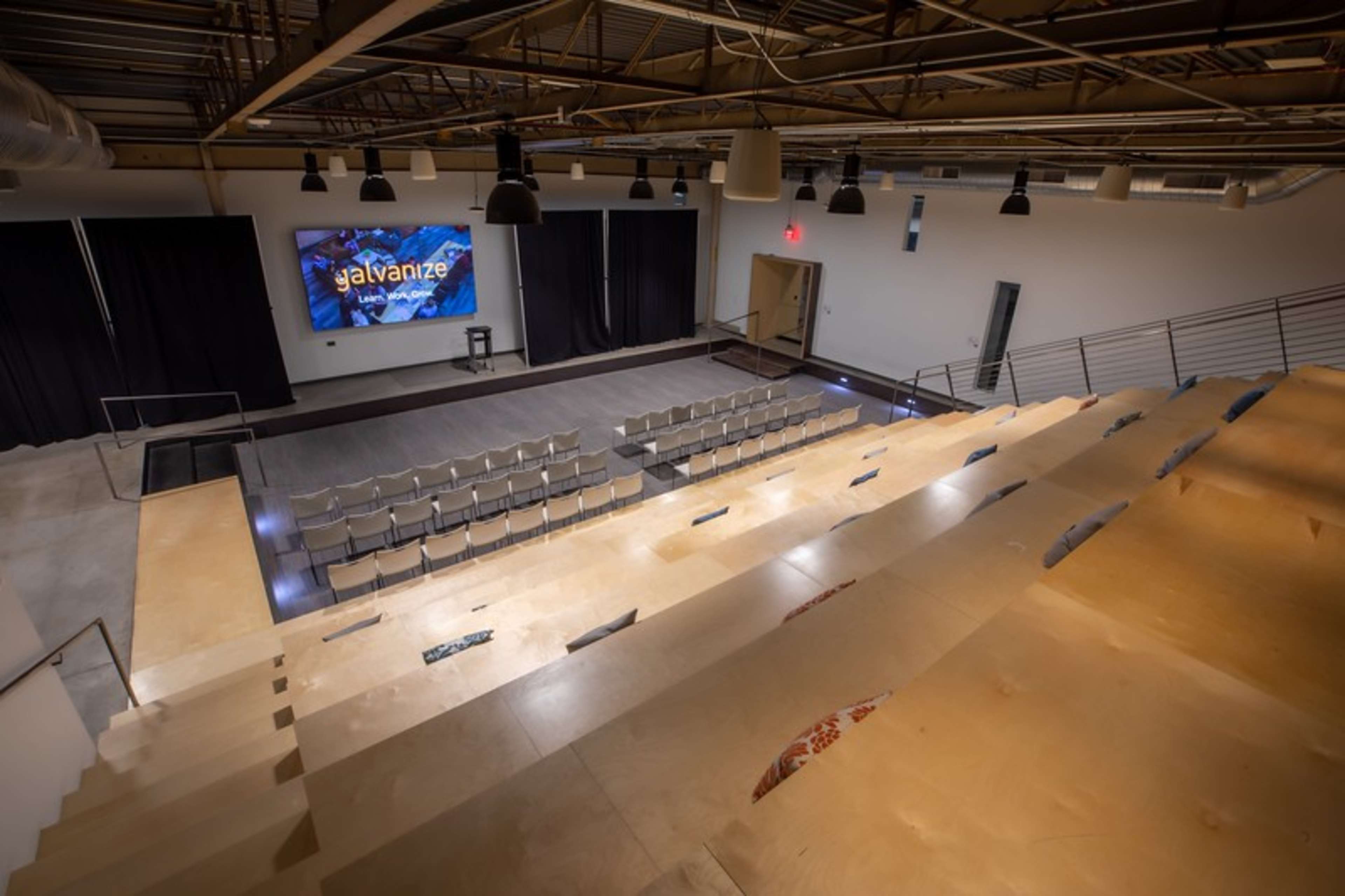 An auditorium with tiered seating, a stage backdrop displaying the word "Galvanize," and a large screen at the front.