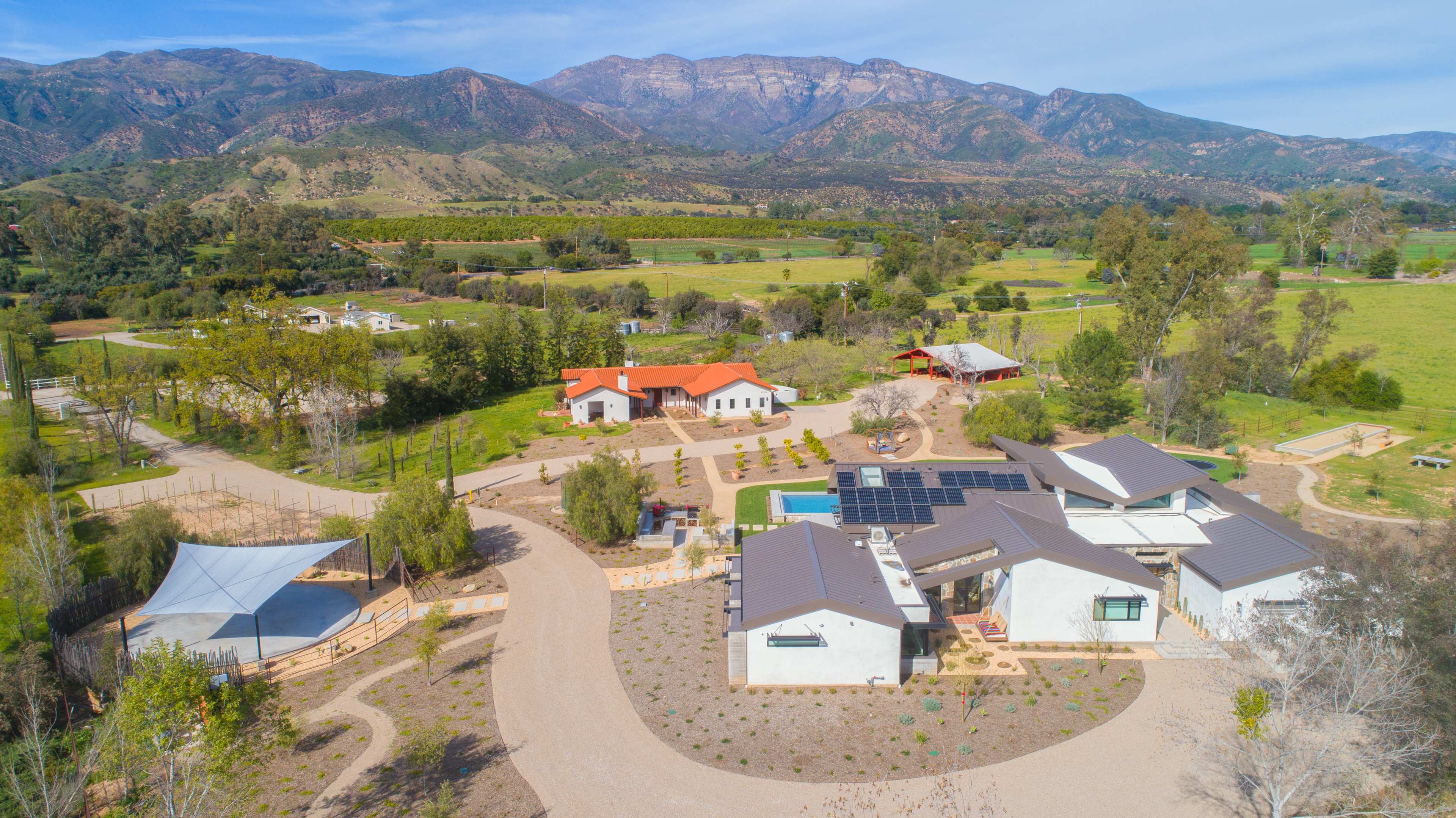 The image shows a sprawling rural property featuring multiple structures, a swimming pool, solar panels, and mountainous terrain in the background.