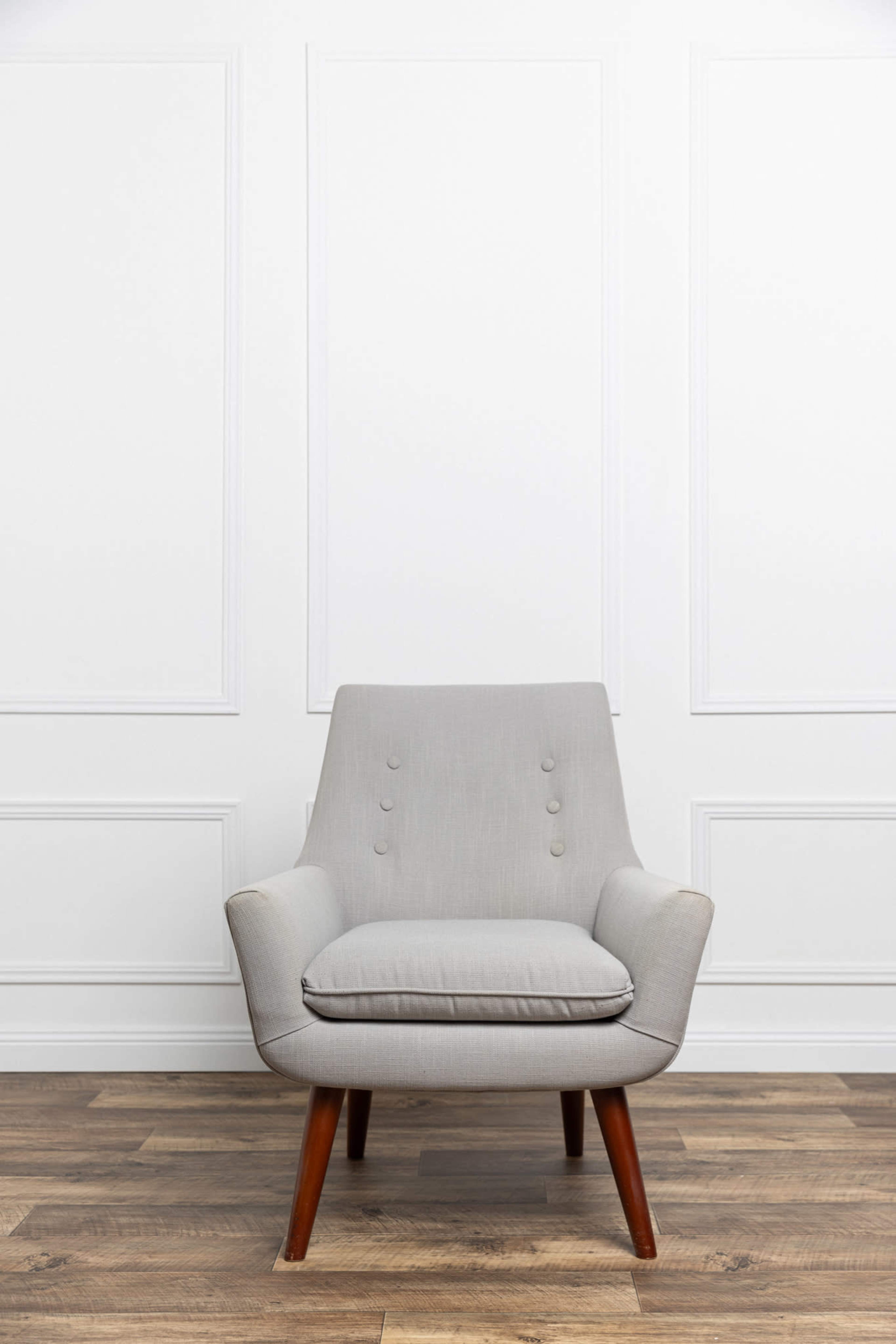 A light gray upholstered chair with wooden legs is positioned in front of a white paneled wall.