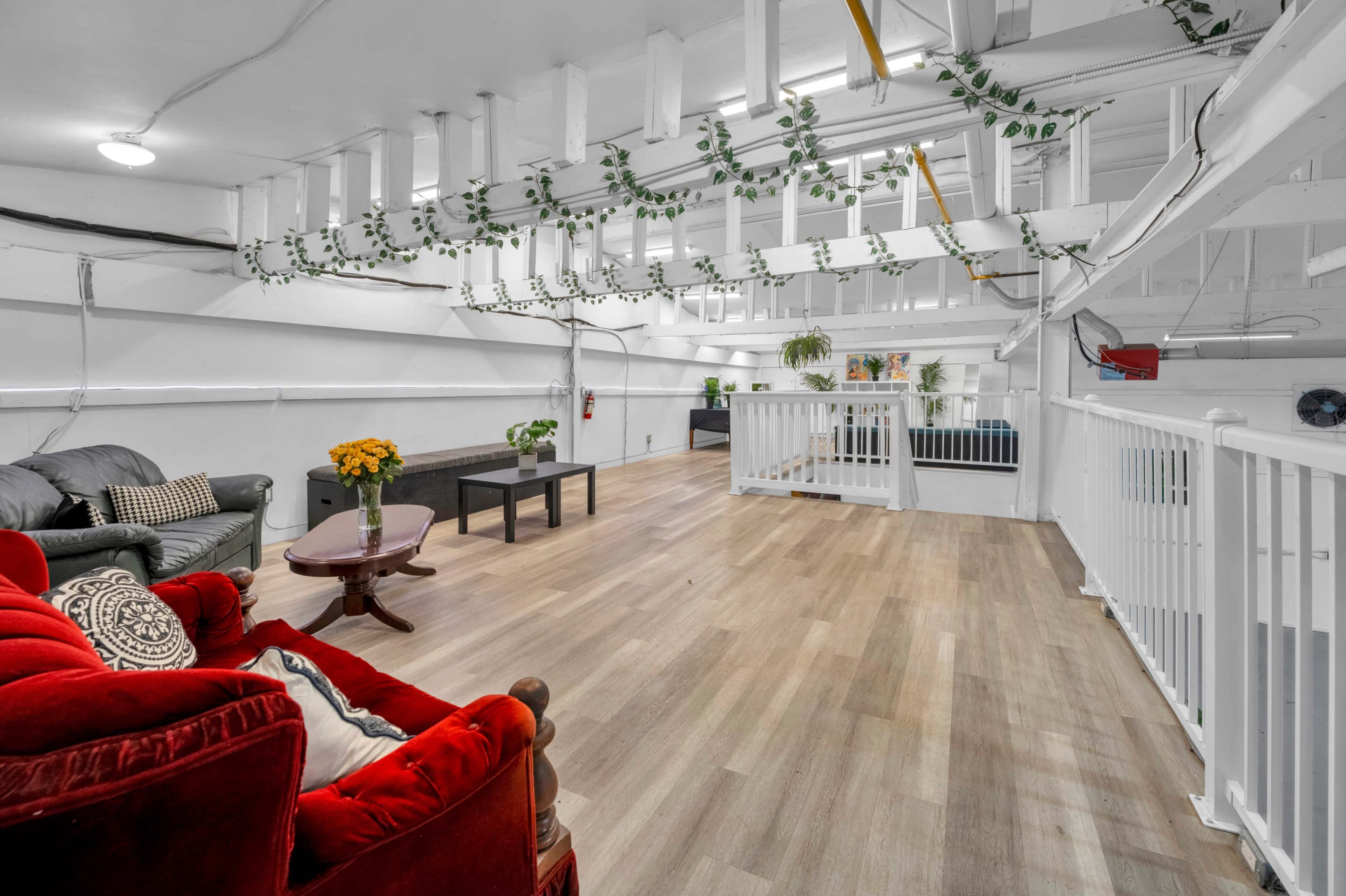 The image shows a spacious loft area with a hardwood floor, featuring a red couch, a small table with a vase of flowers, and various plants along the overhead beams.