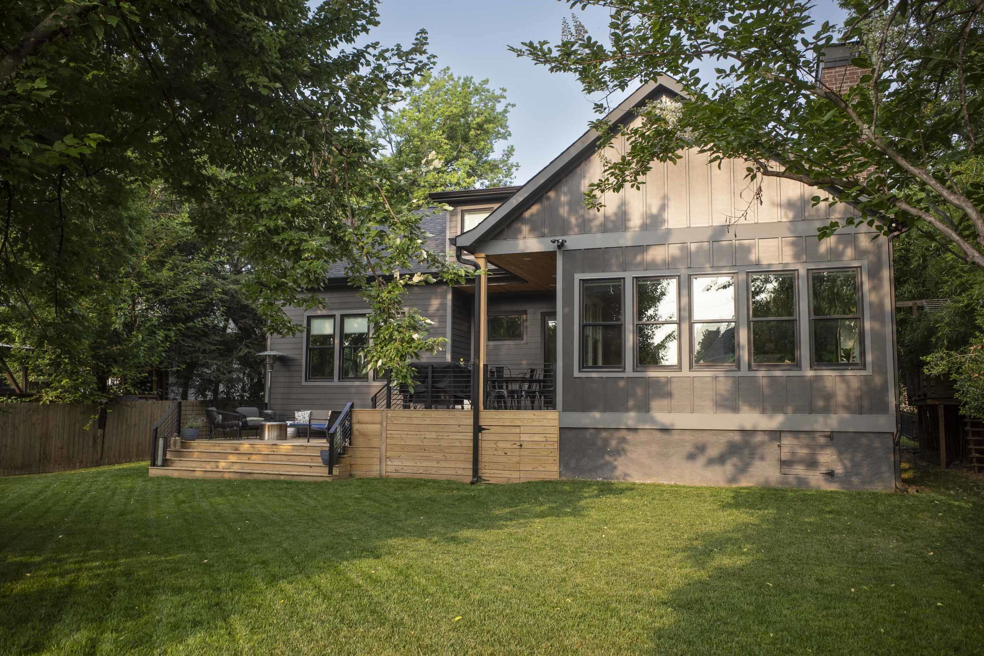 A two-story gray house with large windows and a wooden deck is set in a green yard surrounded by trees.