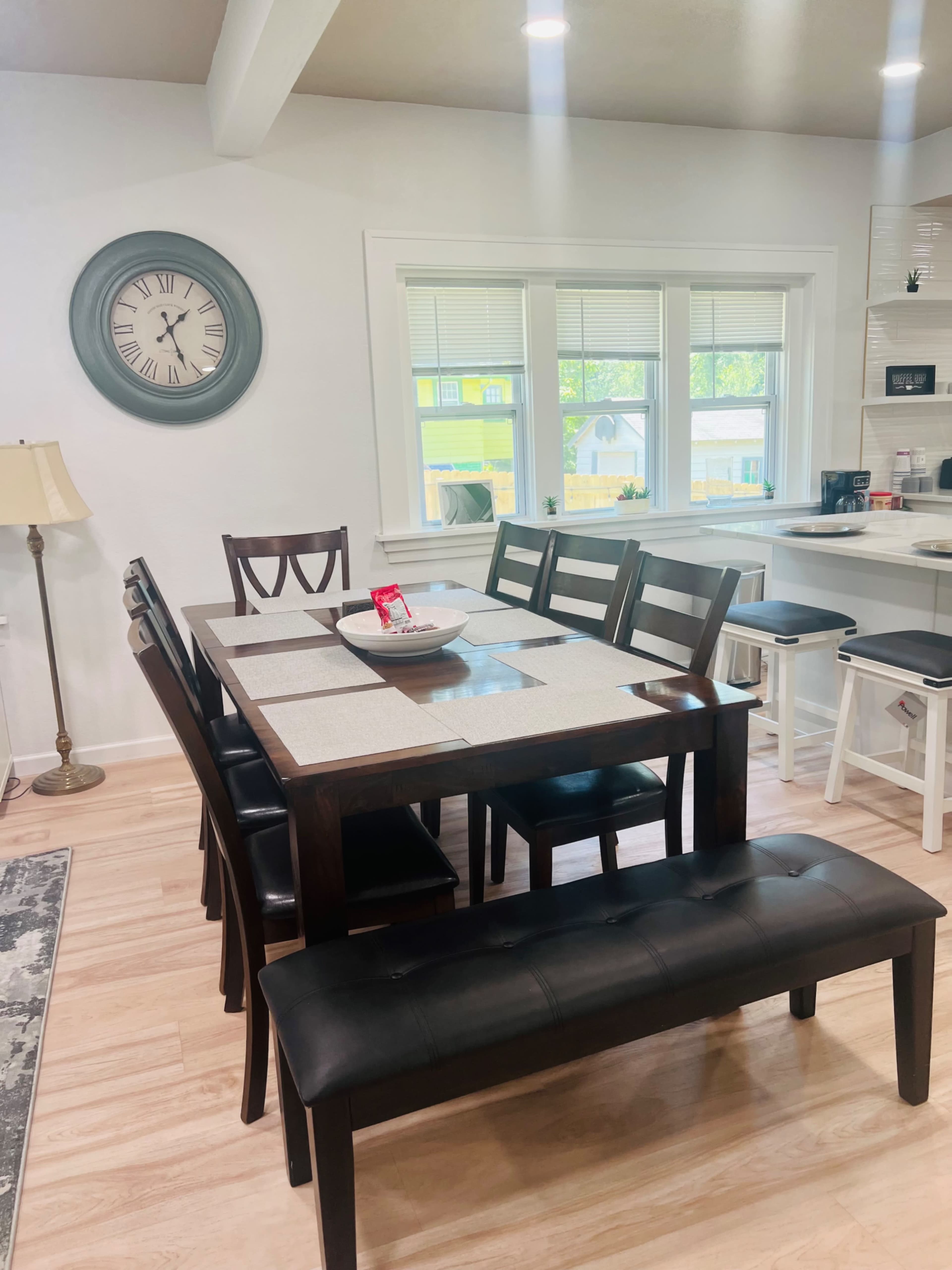 A dining area features a rectangular table surrounded by several chairs and a bench, with a clock on the wall and a window providing natural light.