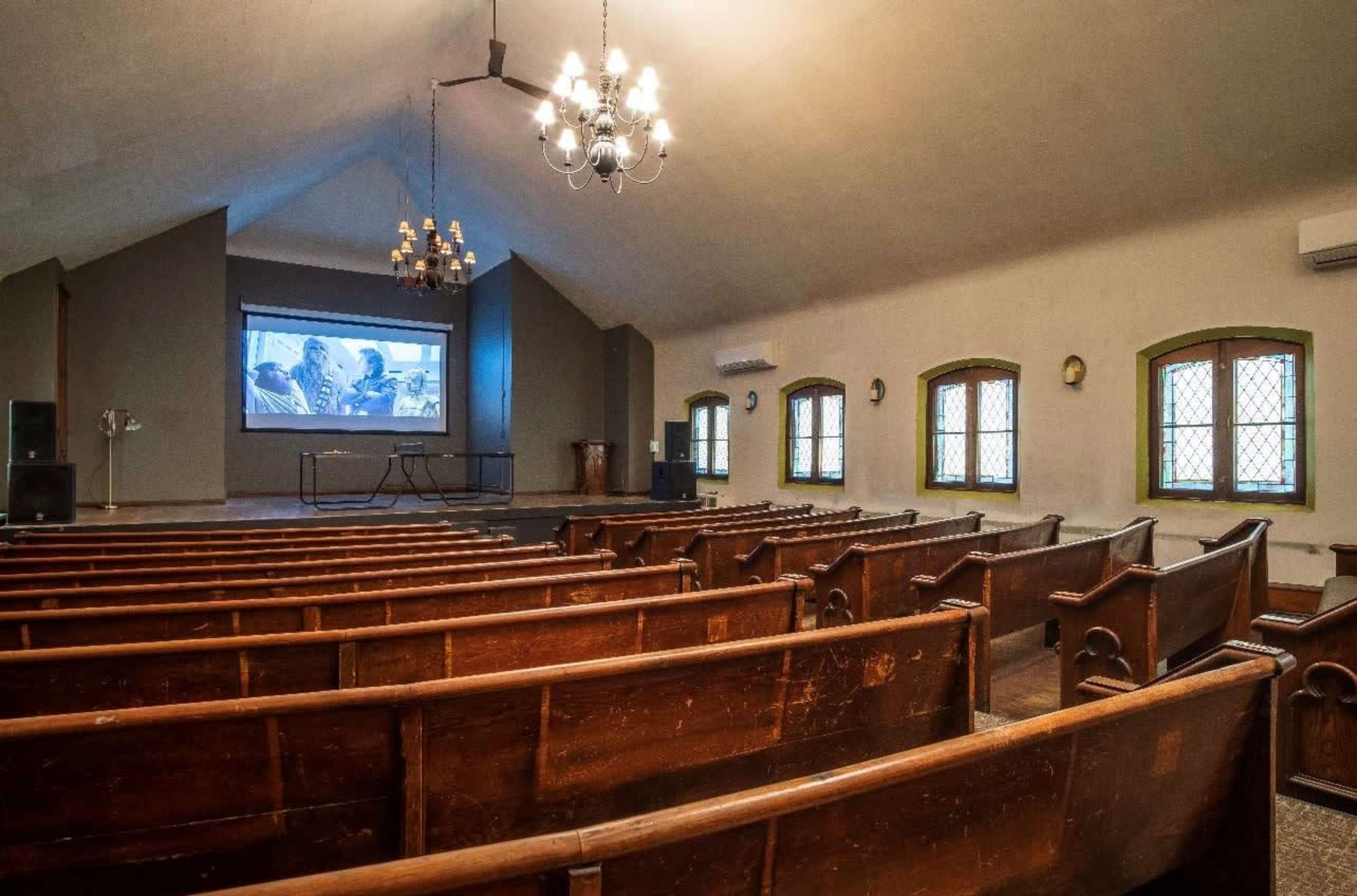 The image shows a sparsely filled auditorium with wooden benches facing a projection screen at the front.