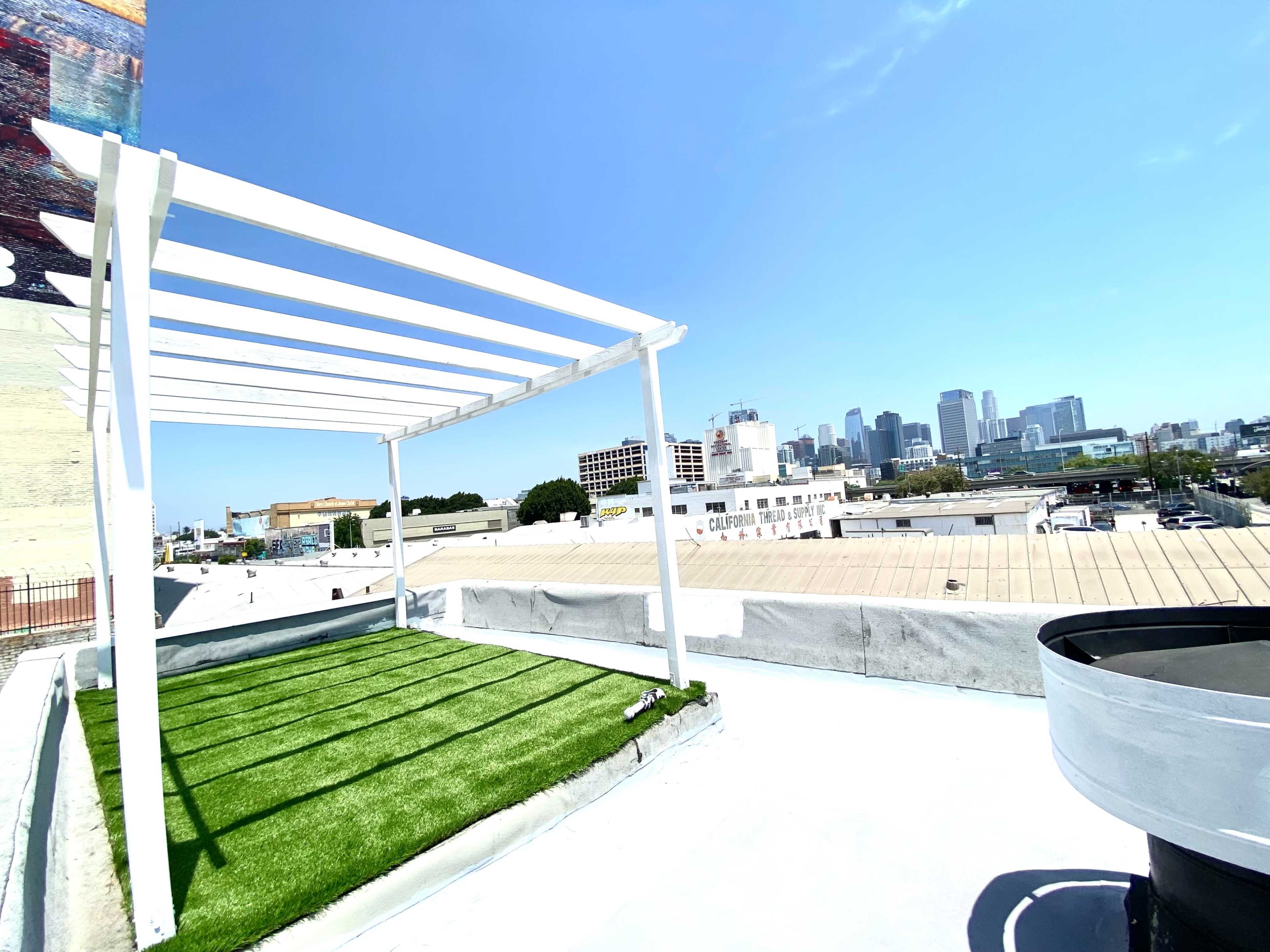 A rooftop terrace with a white pergola, artificial grass, and a view of a city skyline.