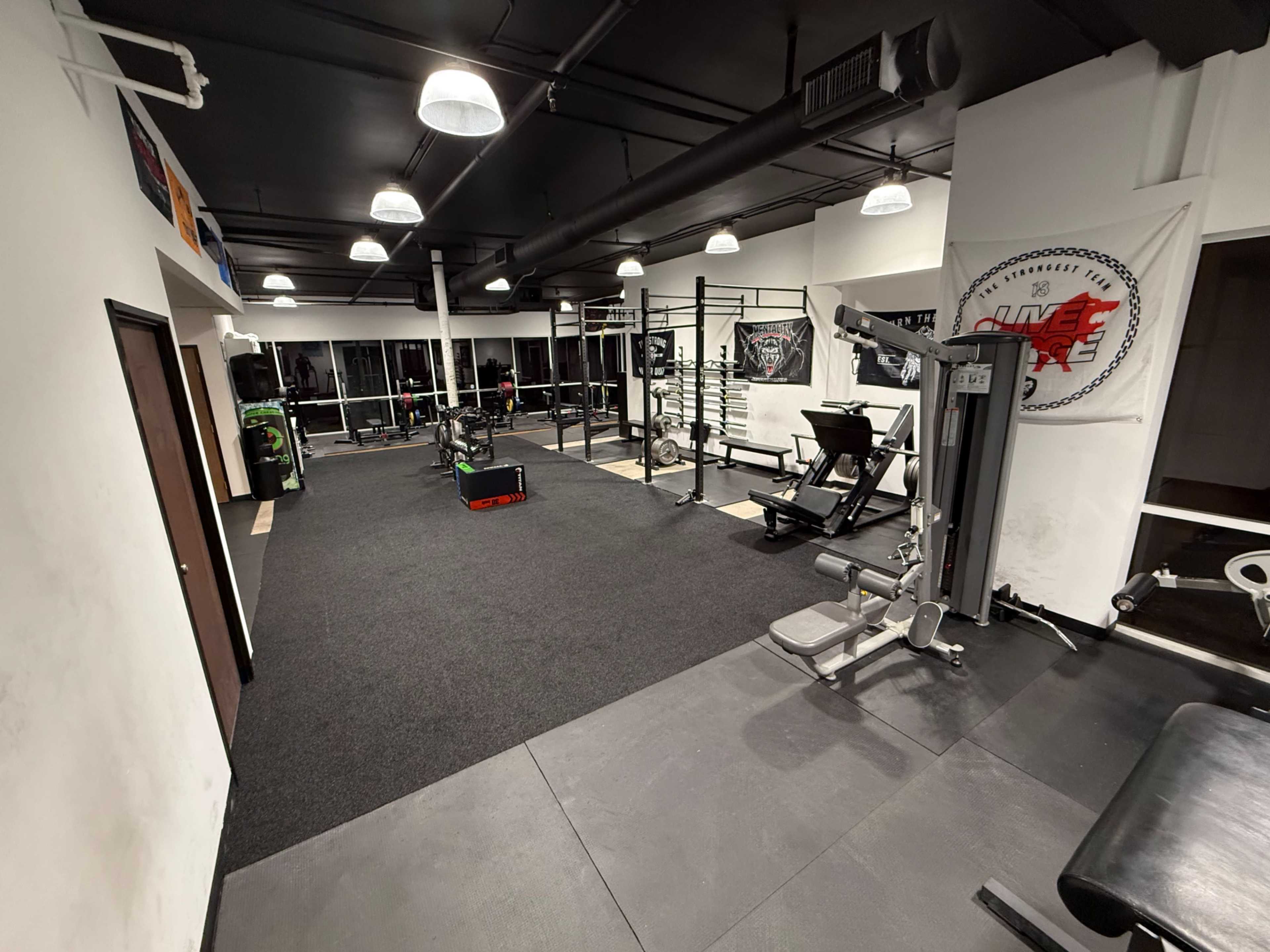 The image shows a well-equipped gym featuring various workout machines, free weights, and open floor space with black rubber flooring.