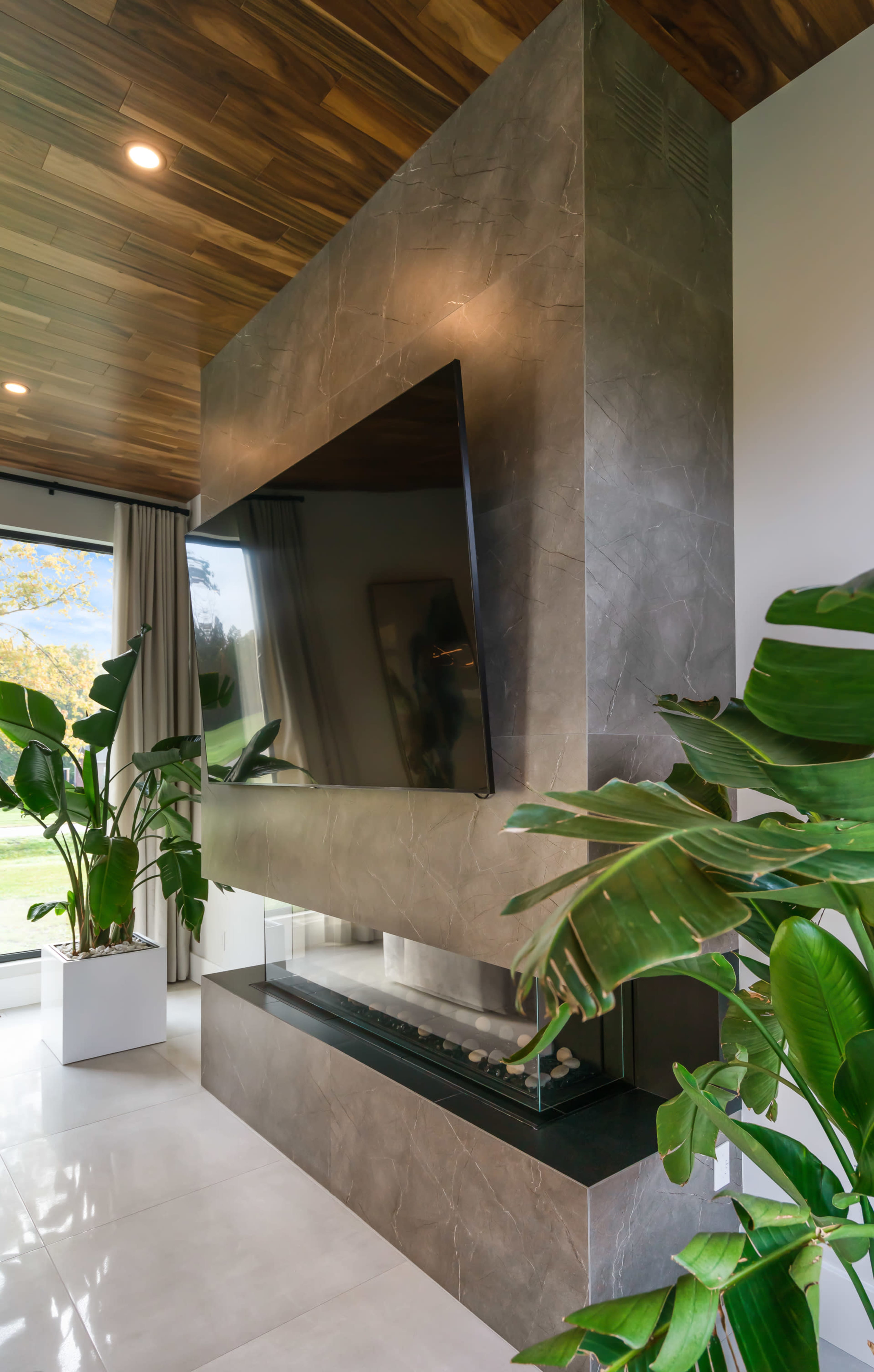 A modern living space featuring a stone fireplace with a mounted flat-screen TV above it and large indoor plants in white containers.