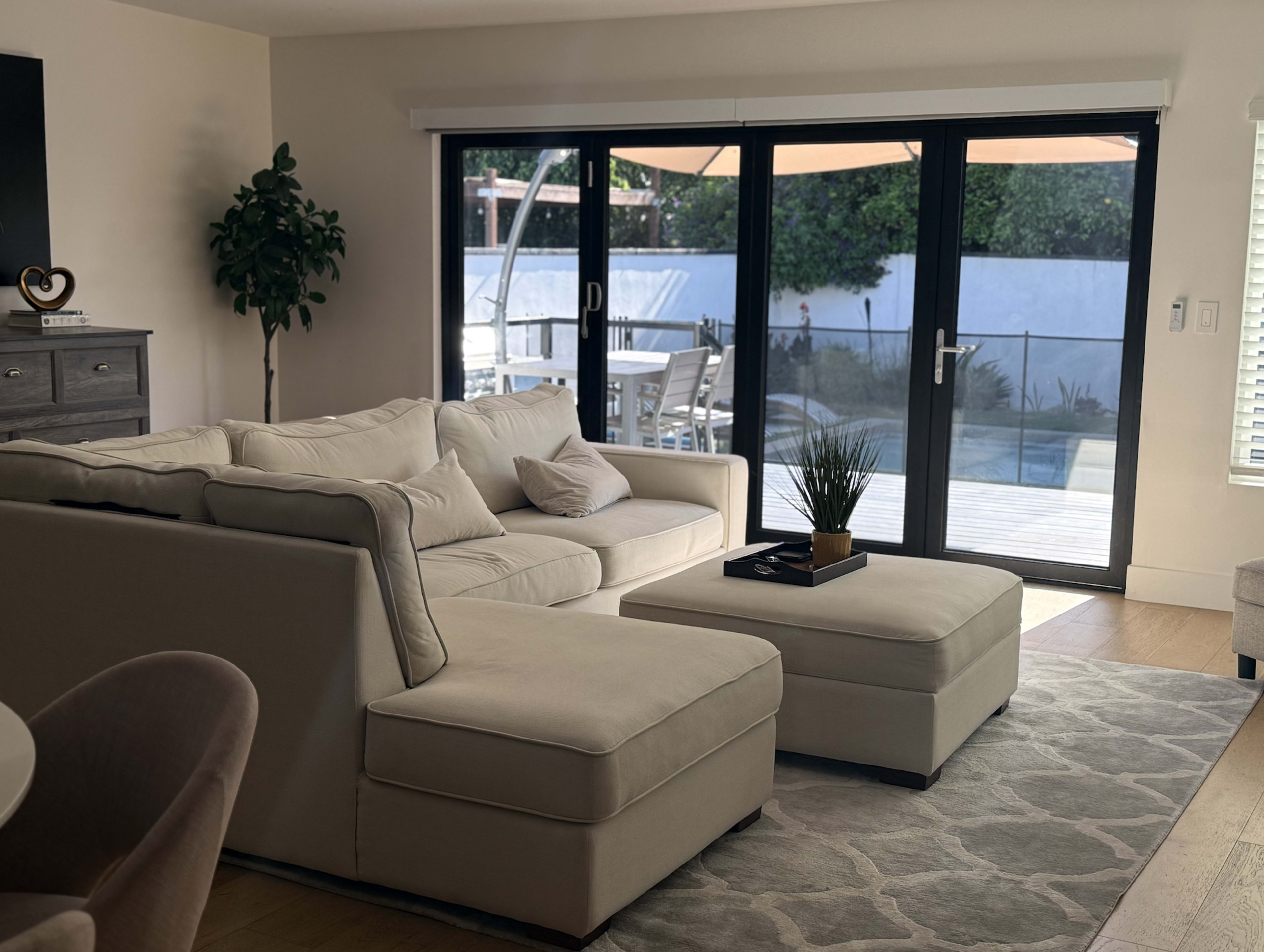 The image shows a light-colored living room with a sectional sofa and an ottoman, leading to large glass doors that open onto an outdoor patio area.