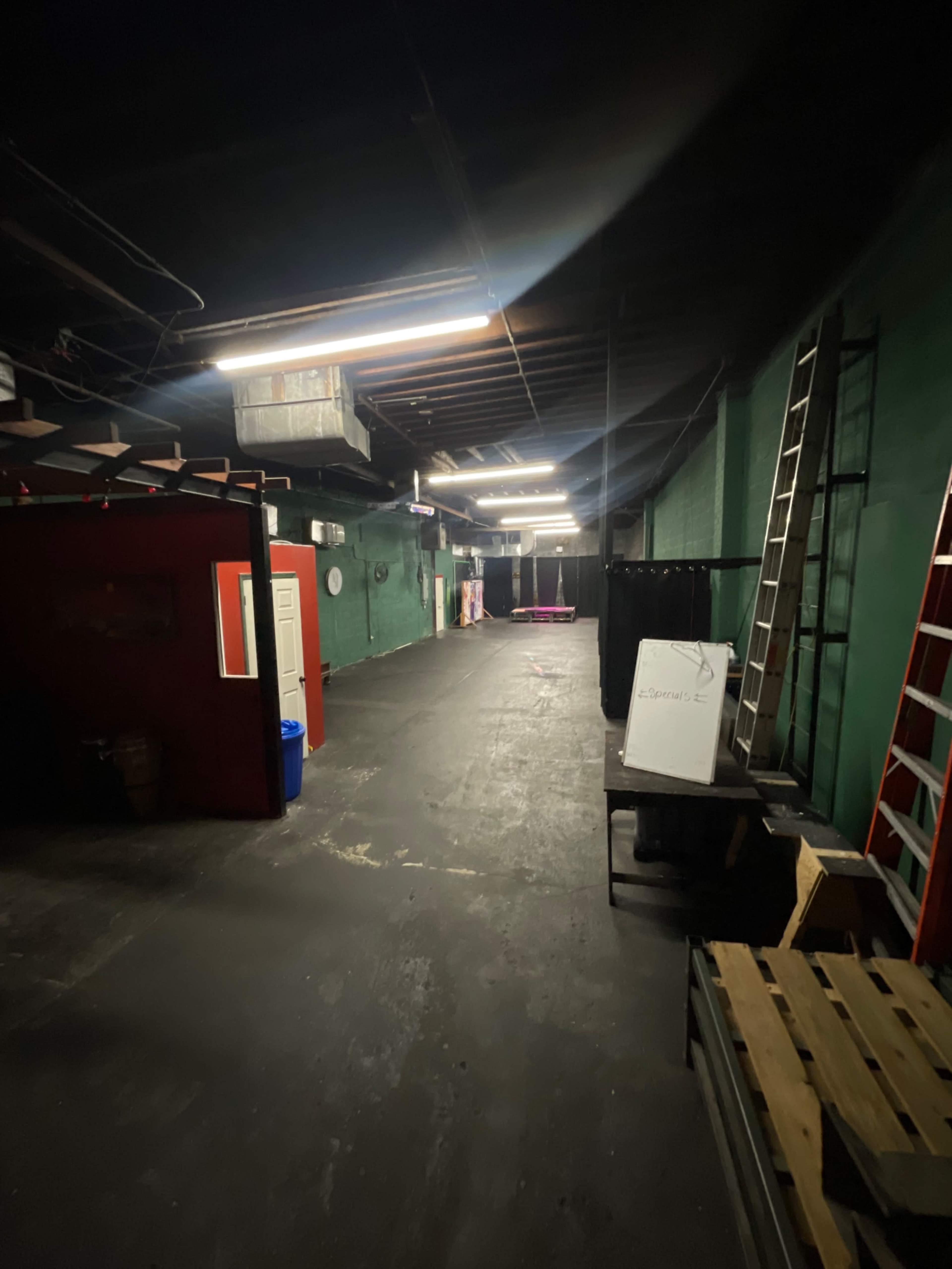 The image shows a dimly lit, industrial-style interior space with green walls, a long hallway, and a mix of ladders and furniture positioned along the sides.